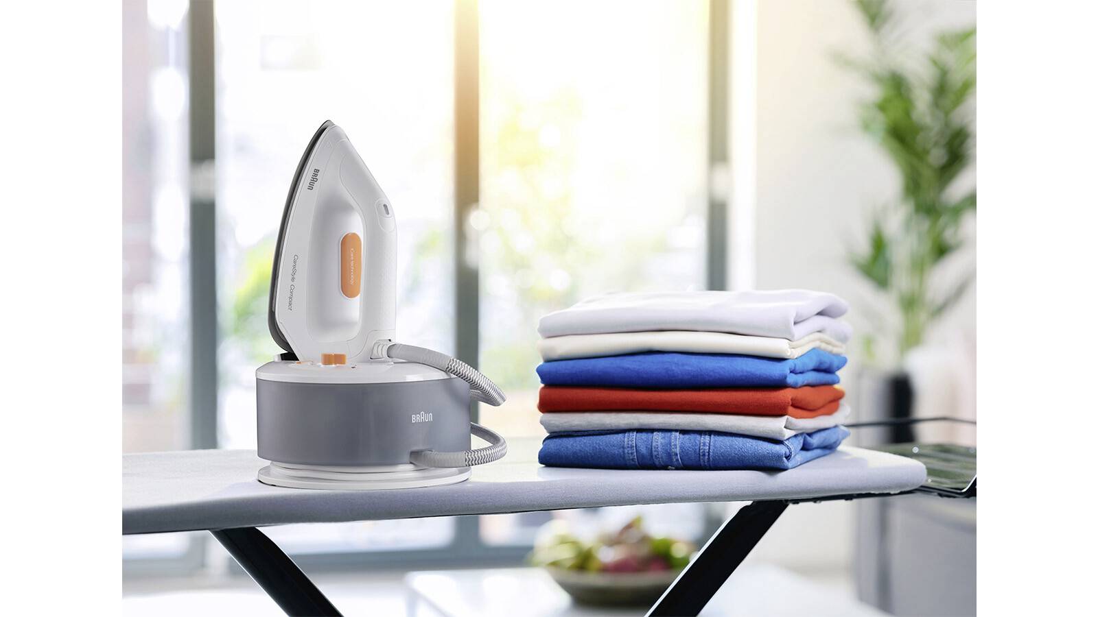 An iron on a board next to a neatly stacked pile of colorful clothes, with a bright window and greenery in the background.