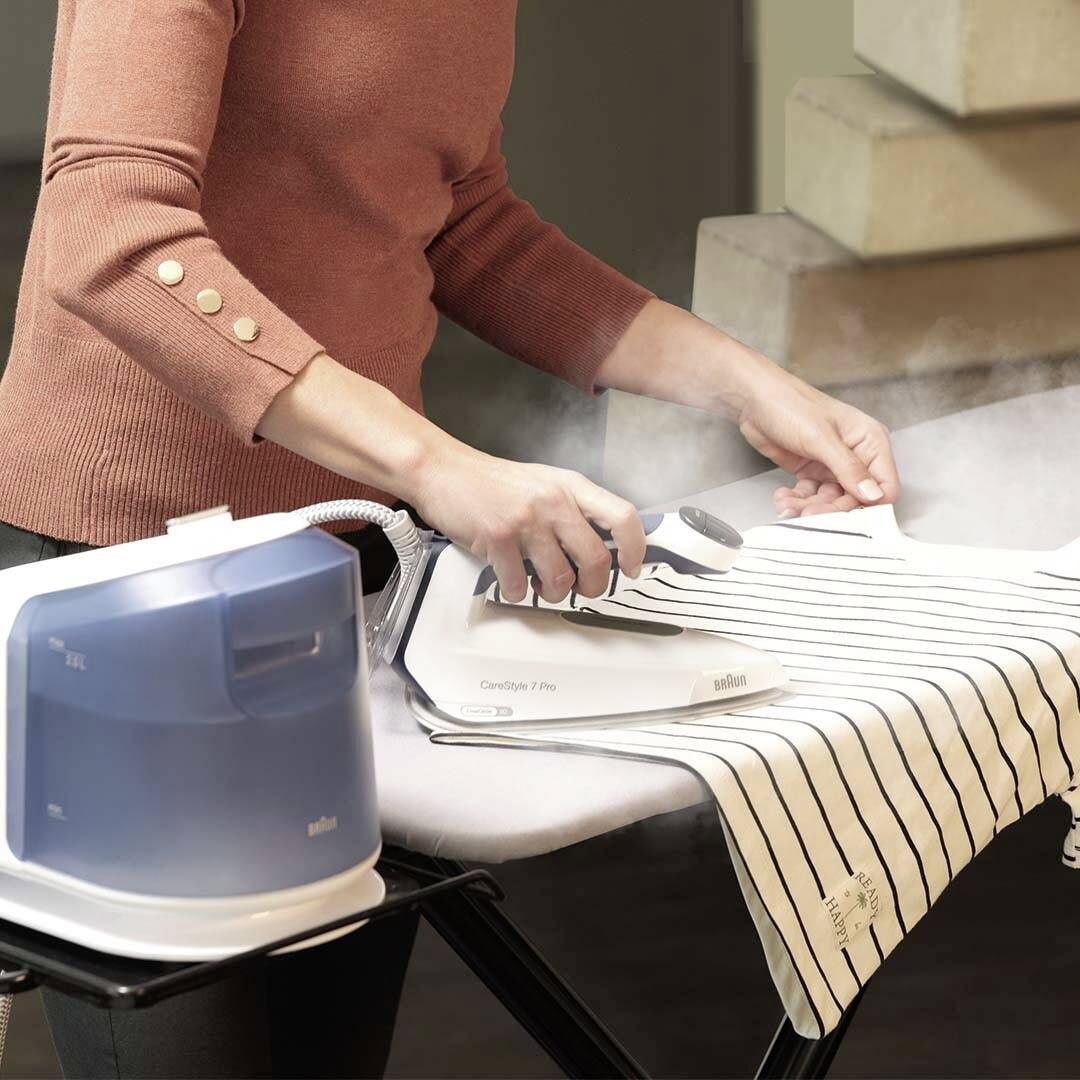 A person is ironing a striped shirt on an ironing board, using a steam iron near a bulky steam generator.