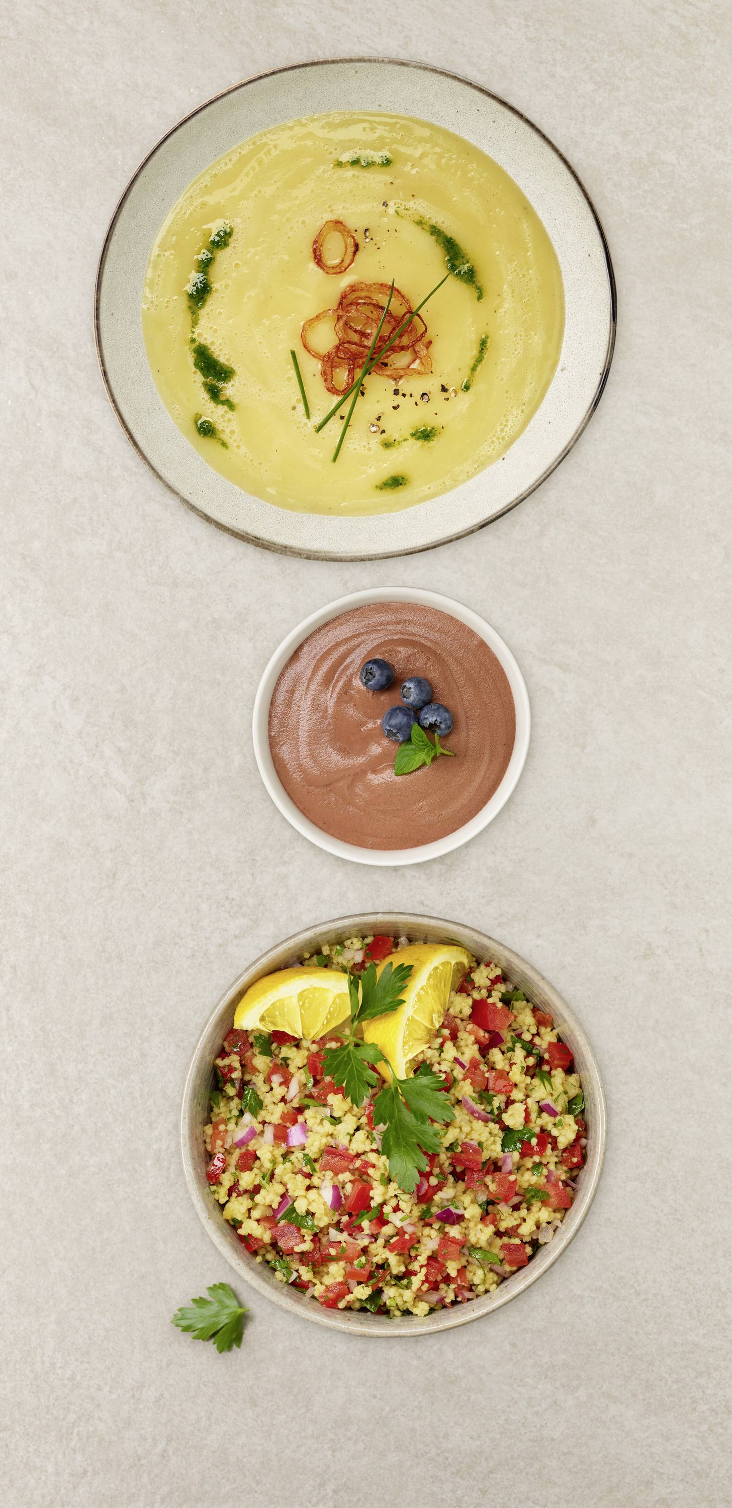 A table displays three dishes: a bowl of yellow soup garnished with herbs, a small bowl of chocolate mousse with blueberries, and a plate of colorful couscous salad with lemon slices.