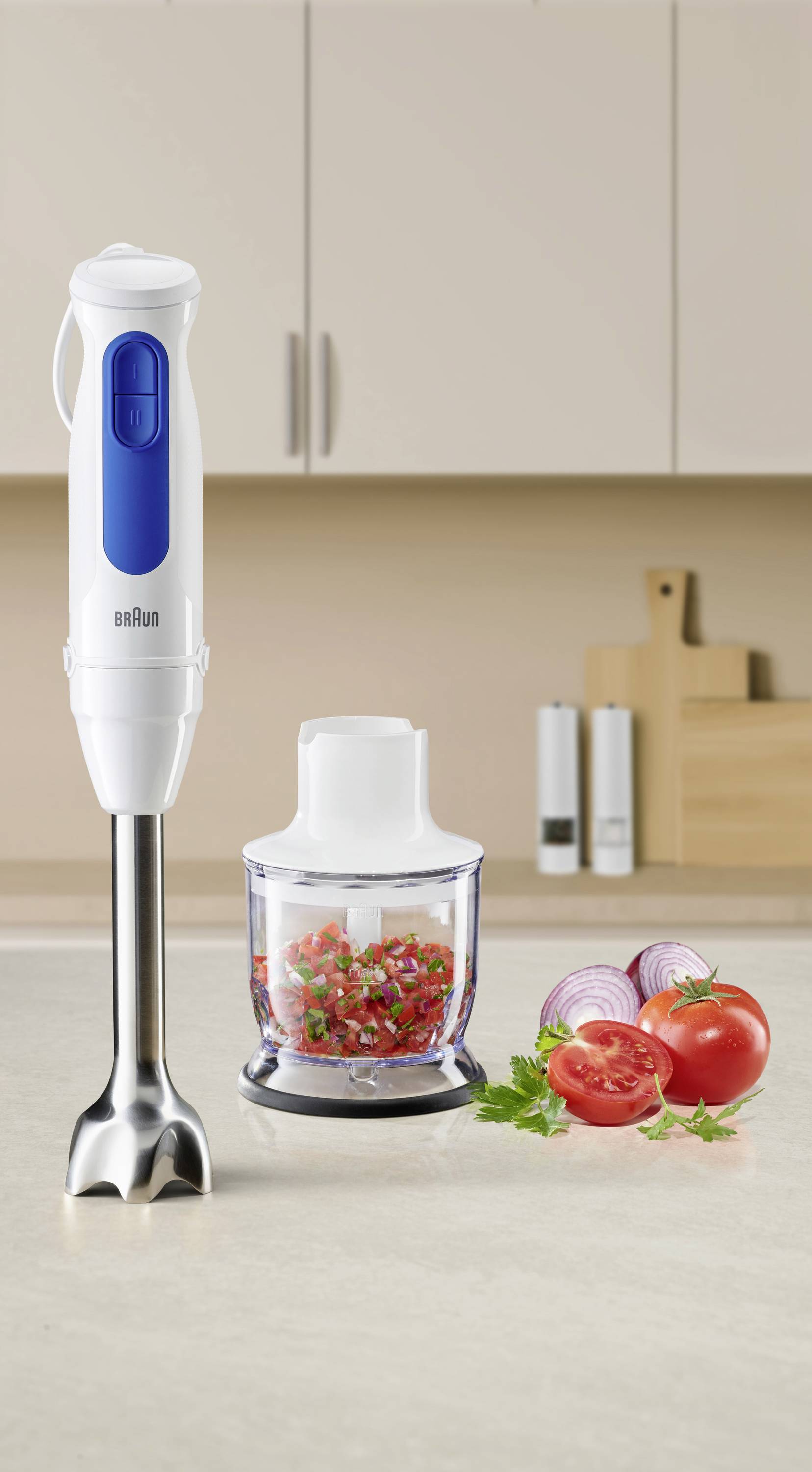 A hand blender set on a kitchen counter with chopped vegetables, including tomatoes and onions, next to it. Kitchen cabinets visible in background.