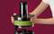 A person placing a blender bowl filled with a green mixture onto a blender base against a maroon background.