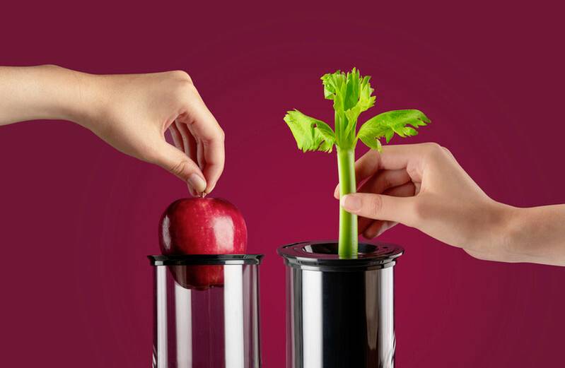 Two hands place a red apple and a celery stalk into separate tall, transparent containers against a solid maroon background.