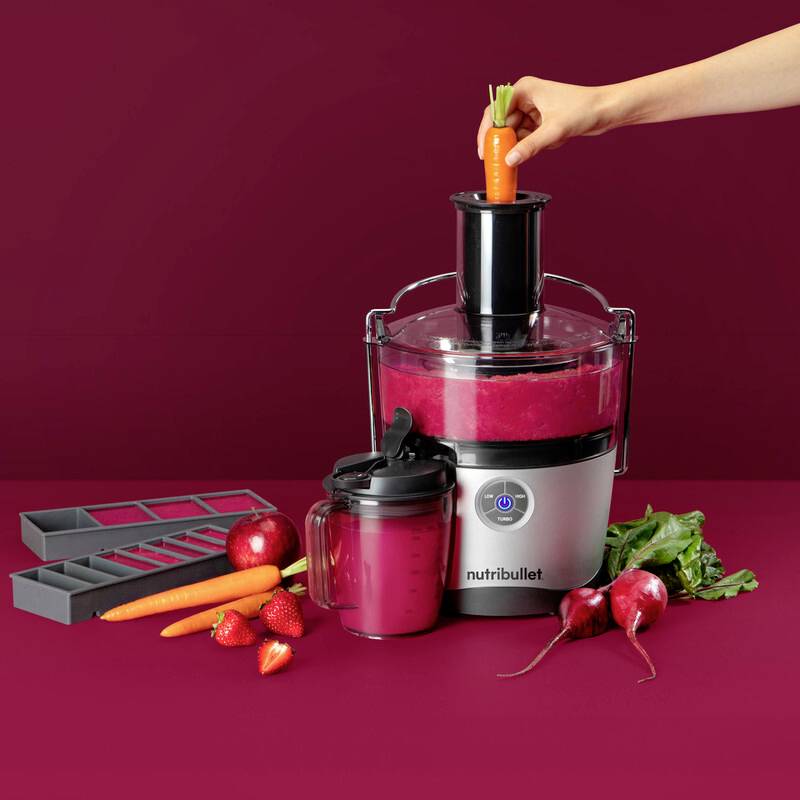A person inserts a carrot into a Nutribullet juicer surrounded by strawberries, carrots, and beets with a finished juice in front.