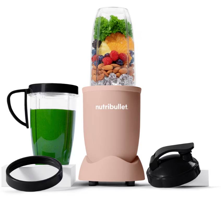A pink NutriBullet blender with fruit, almonds, and kale inside the cup. Nearby are a green drink in a to-go cup, a lid, and a seal ring.