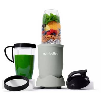 A NutriBullet blender filled with assorted fruits, vegetables, and nuts beside a green drink in a cup and a black lid.