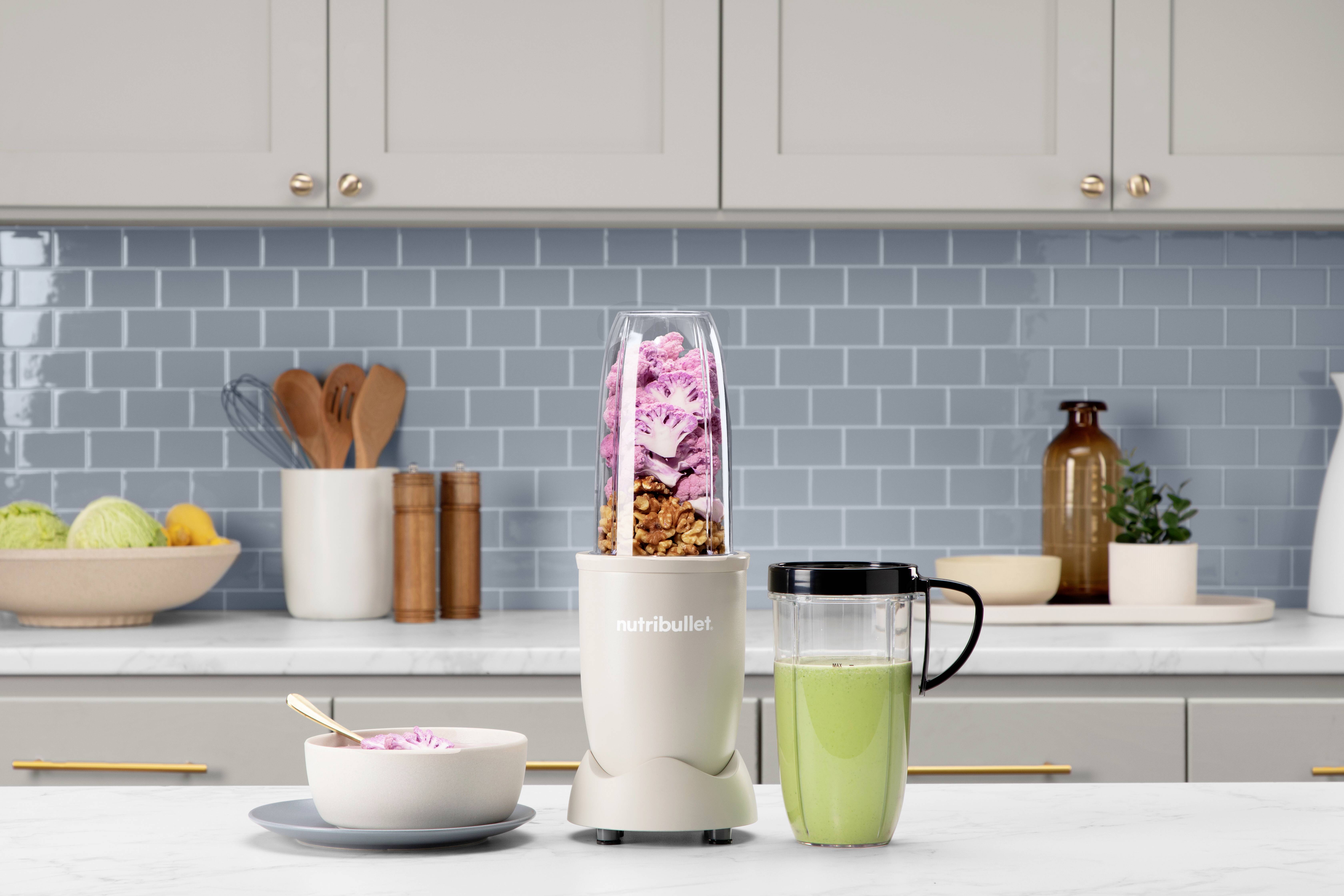A NutriBullet blender sits on a kitchen counter, filled with pink smoothie ingredients and nuts. Beside it is a glass with green smoothie.