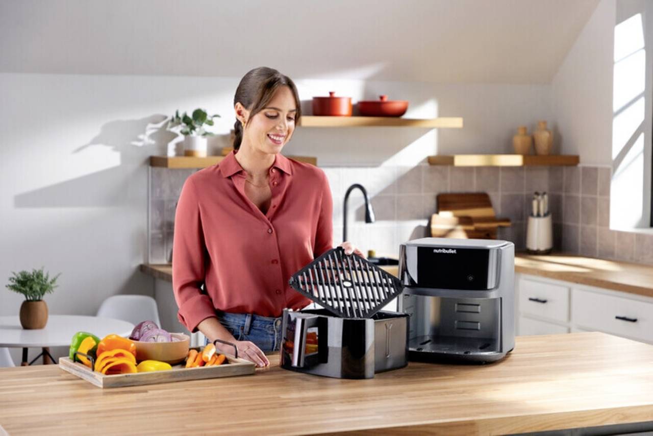 A person in a kitchen places a rack into an air fryer on a counter with cut vegetables. The kitchen has modern decor and potted plants.