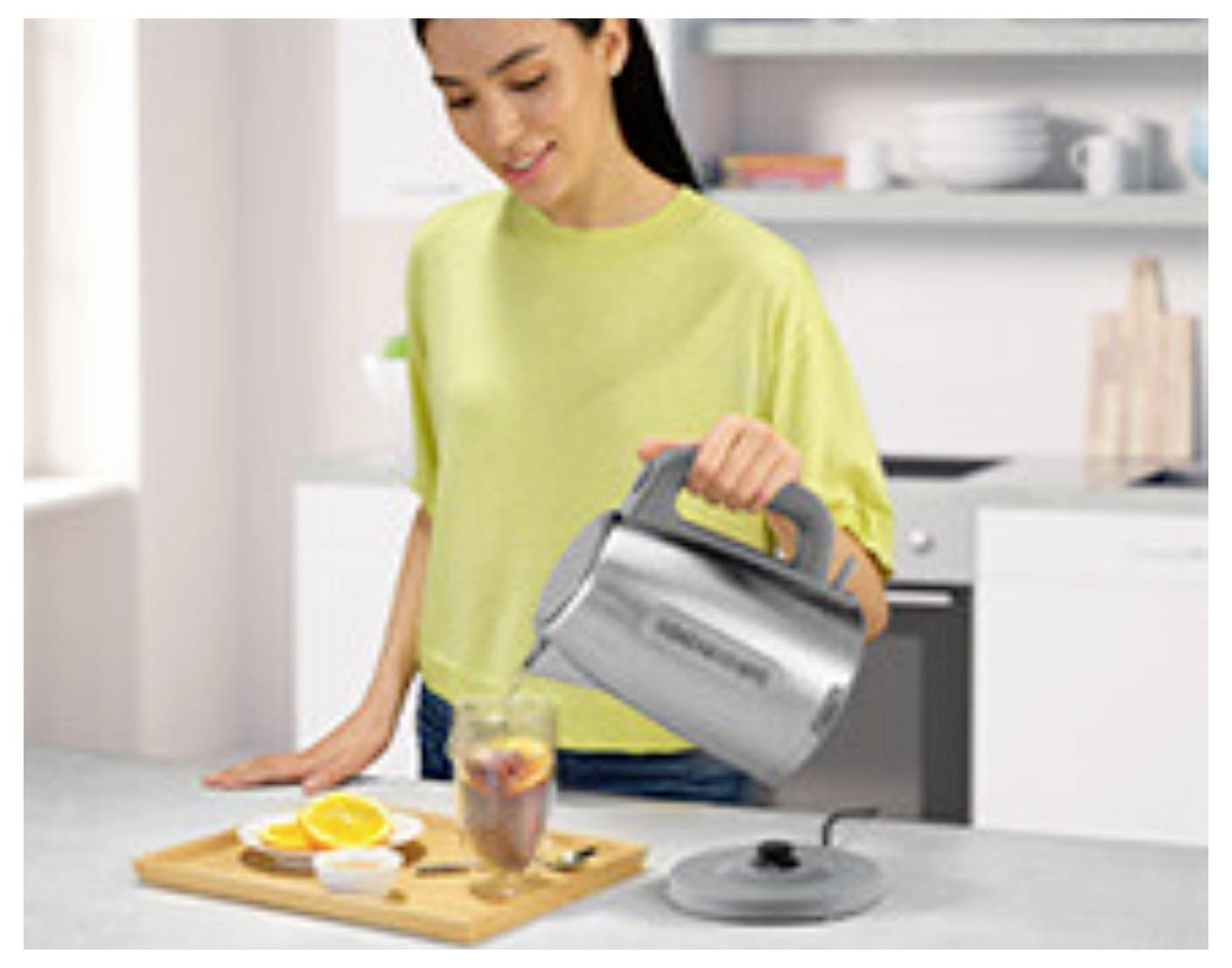A person in a kitchen pours hot water from an electric kettle into a glass with tea. A cutting board with sliced oranges and tea bags is nearby.