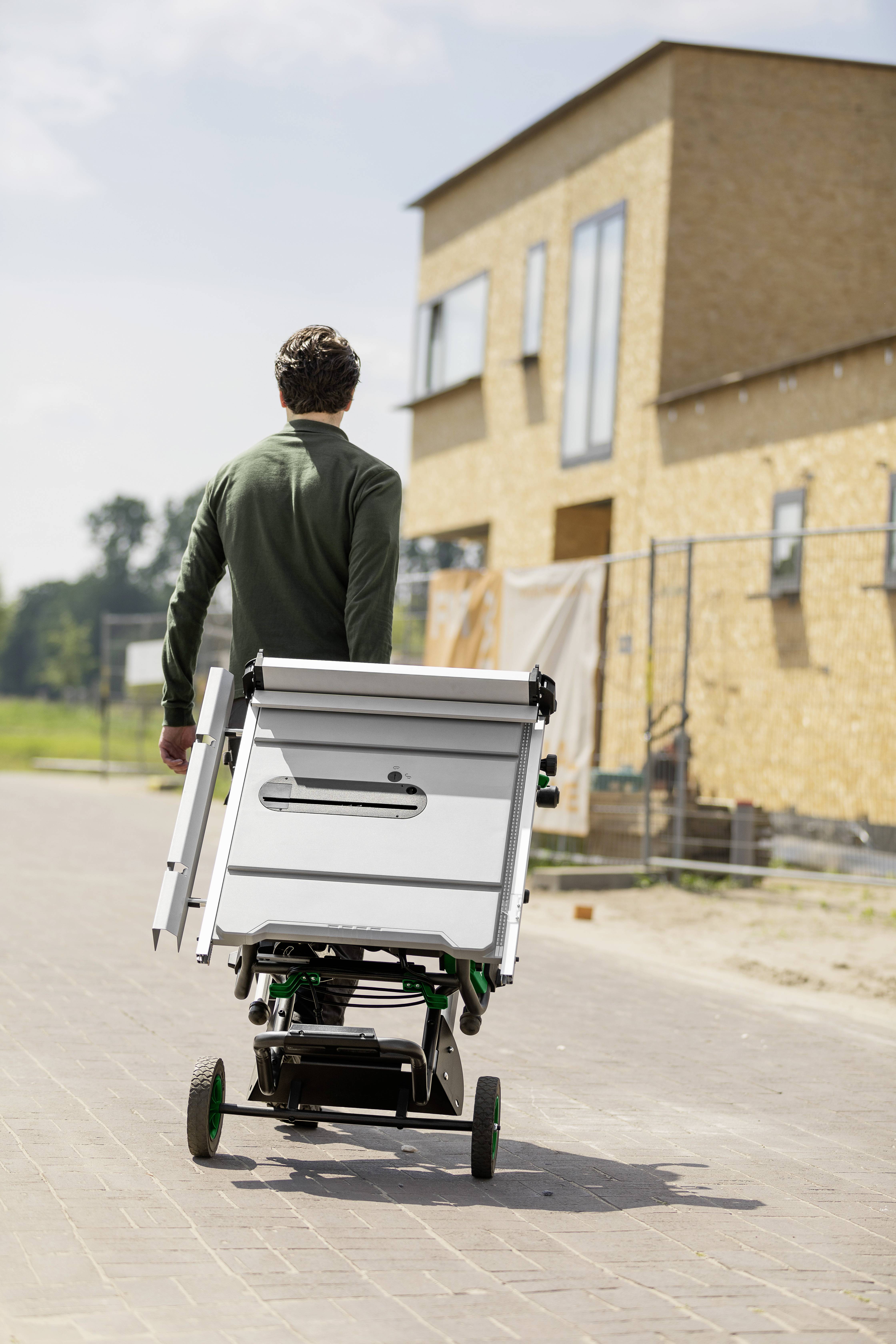 A person walks away from the camera, pushing a mobile cart on a paved path. A large building under construction is in the background.