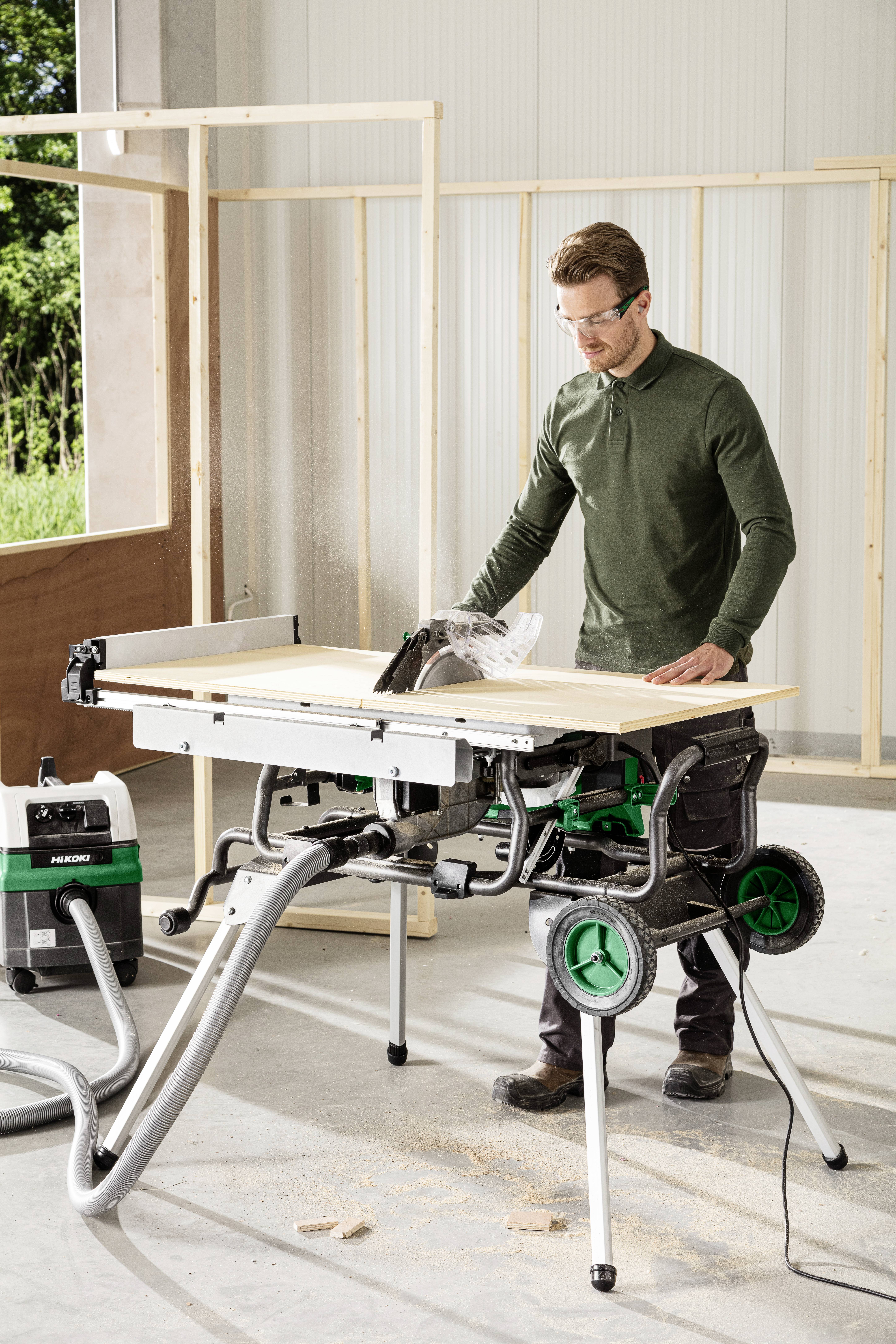 A person in safety glasses operates a table saw in a workshop, cutting a piece of wood. Construction frames and tools surround them.
