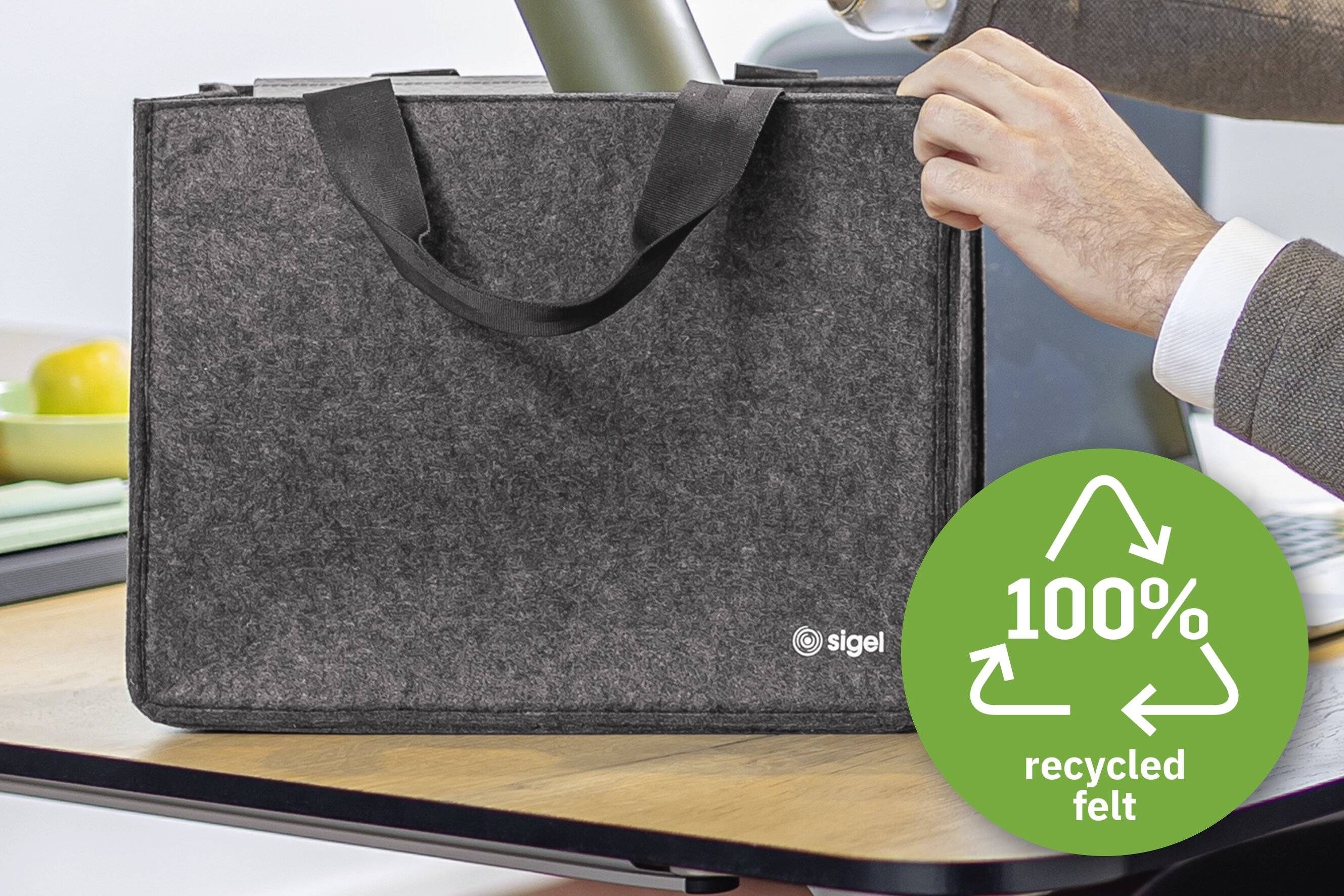 A person holding a gray felt bag on a table. The bag has two handles and a logo. A green badge says '100% recycled felt' with arrows indicating recycling.