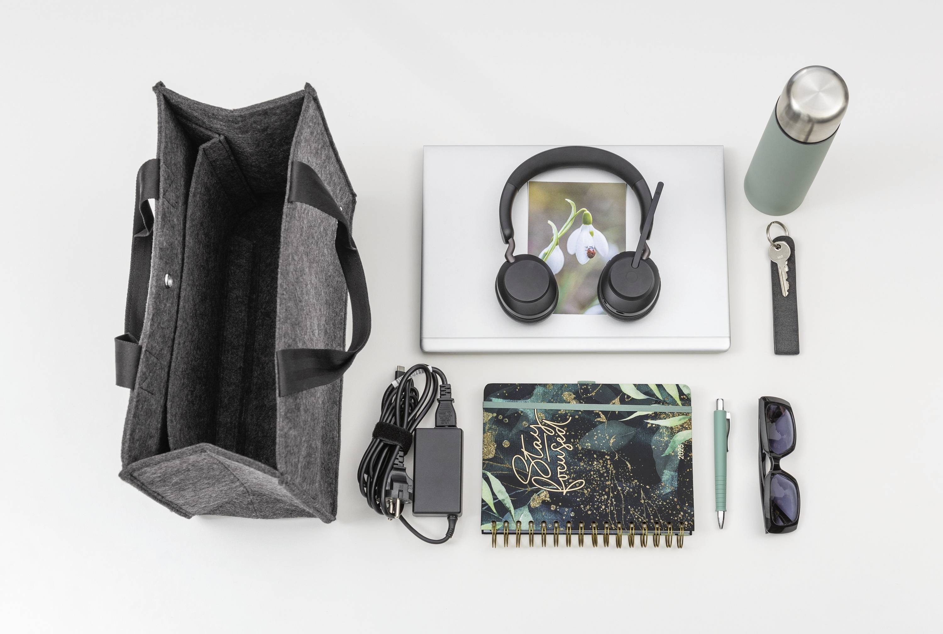 A neatly arranged collection of daily essentials: a gray tote bag, laptop, headphones, notebook, water bottle, charger, sunglasses, pen, and keychain on a white surface.
