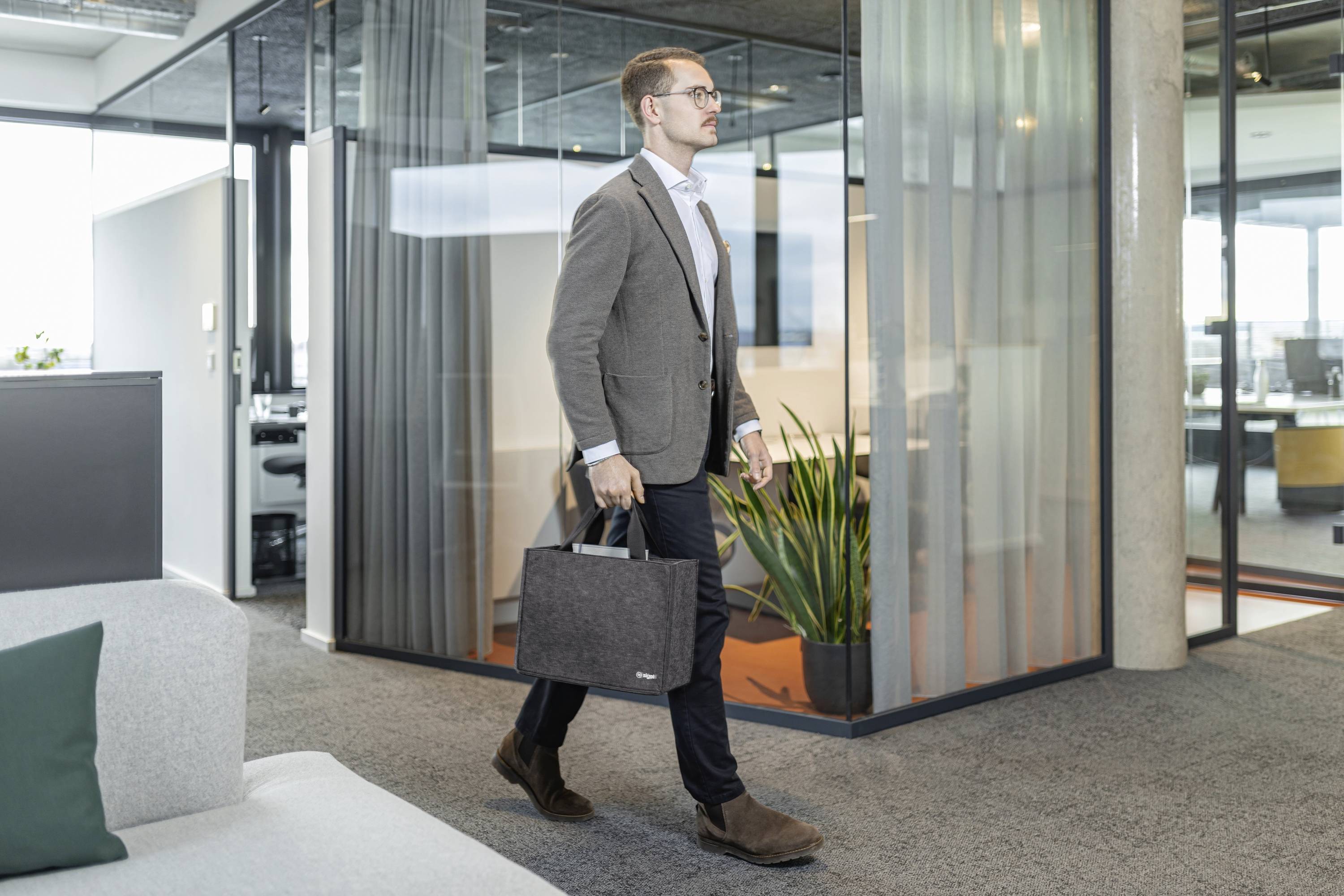 A man in business attire walks through a modern office with glass partitions, carrying a laptop bag, conveying a professional environment.