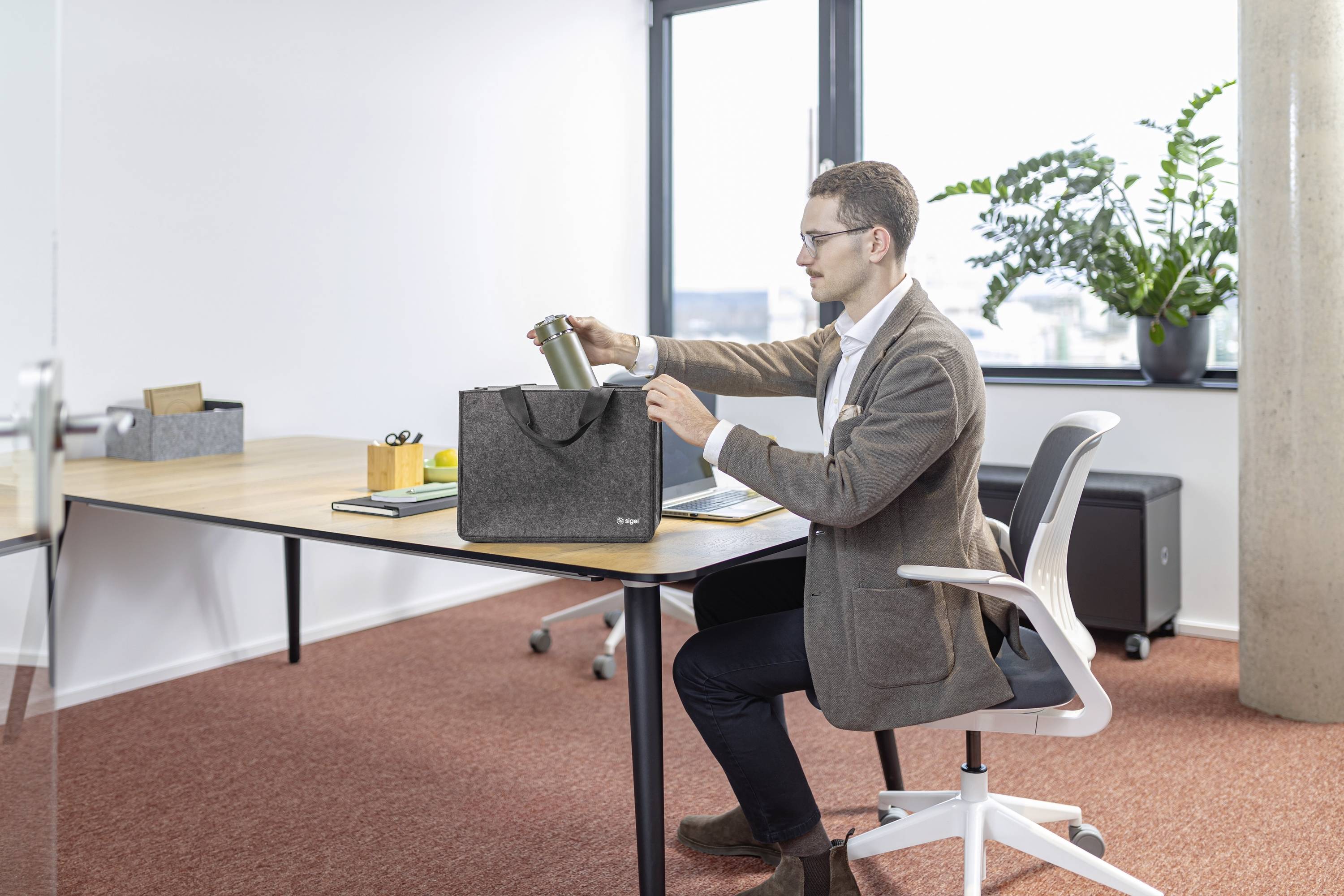 A person sits at a desk in an office, placing a thermos into a gray bag. The room has a large window, potted plant, and office equipment.