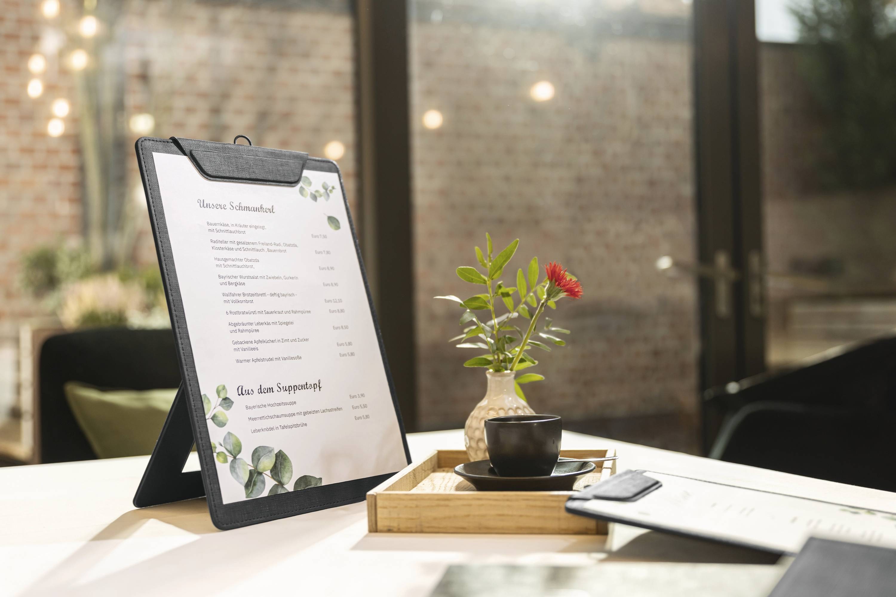 A clipboard holds a menu on a table beside a wooden tray with a small potted plant. There's a soft focus of a bright room in the background.