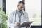 A doctor in a white coat and stethoscope reviews notes on a clipboard in a bright office with a plant and window in the background.