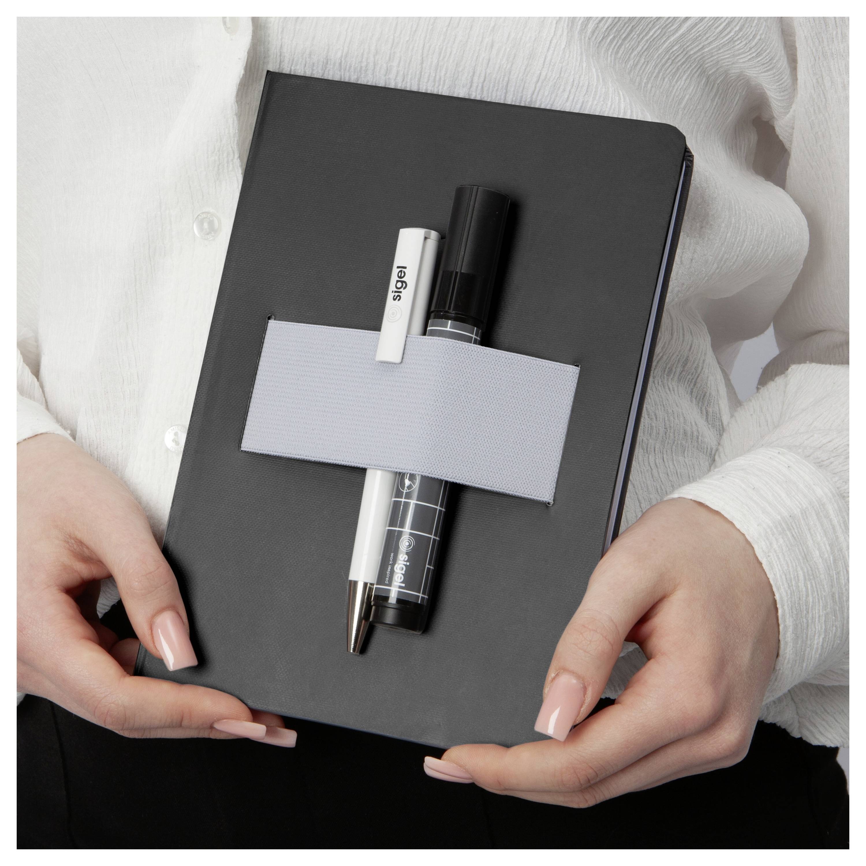 Person holding a black notebook with a pen and white marker strapped across the front by a gray band.