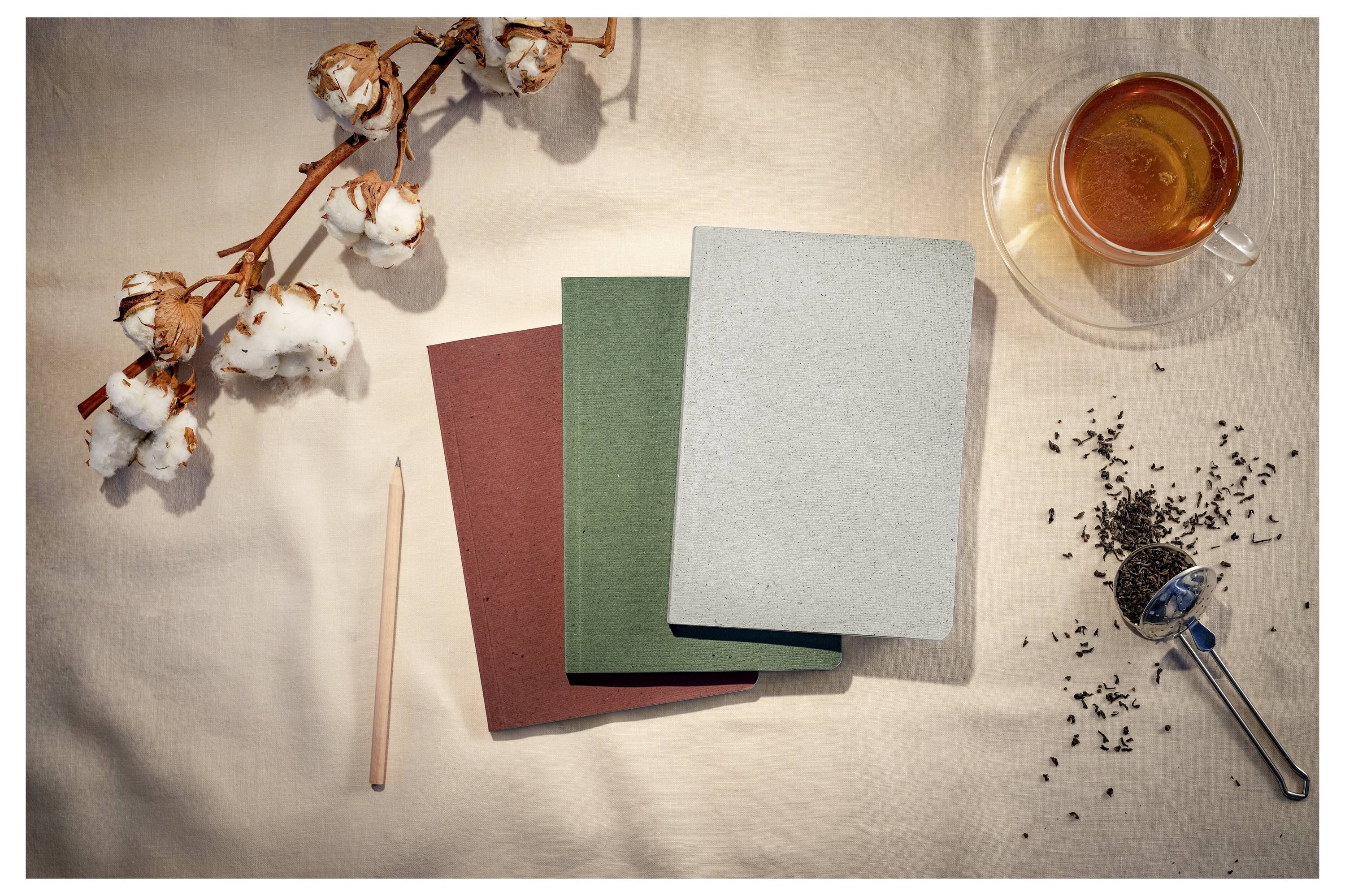 Three blank notebooks with a pencil, cotton stem, teacup, and loose tea leaves arranged on a beige tabletop.