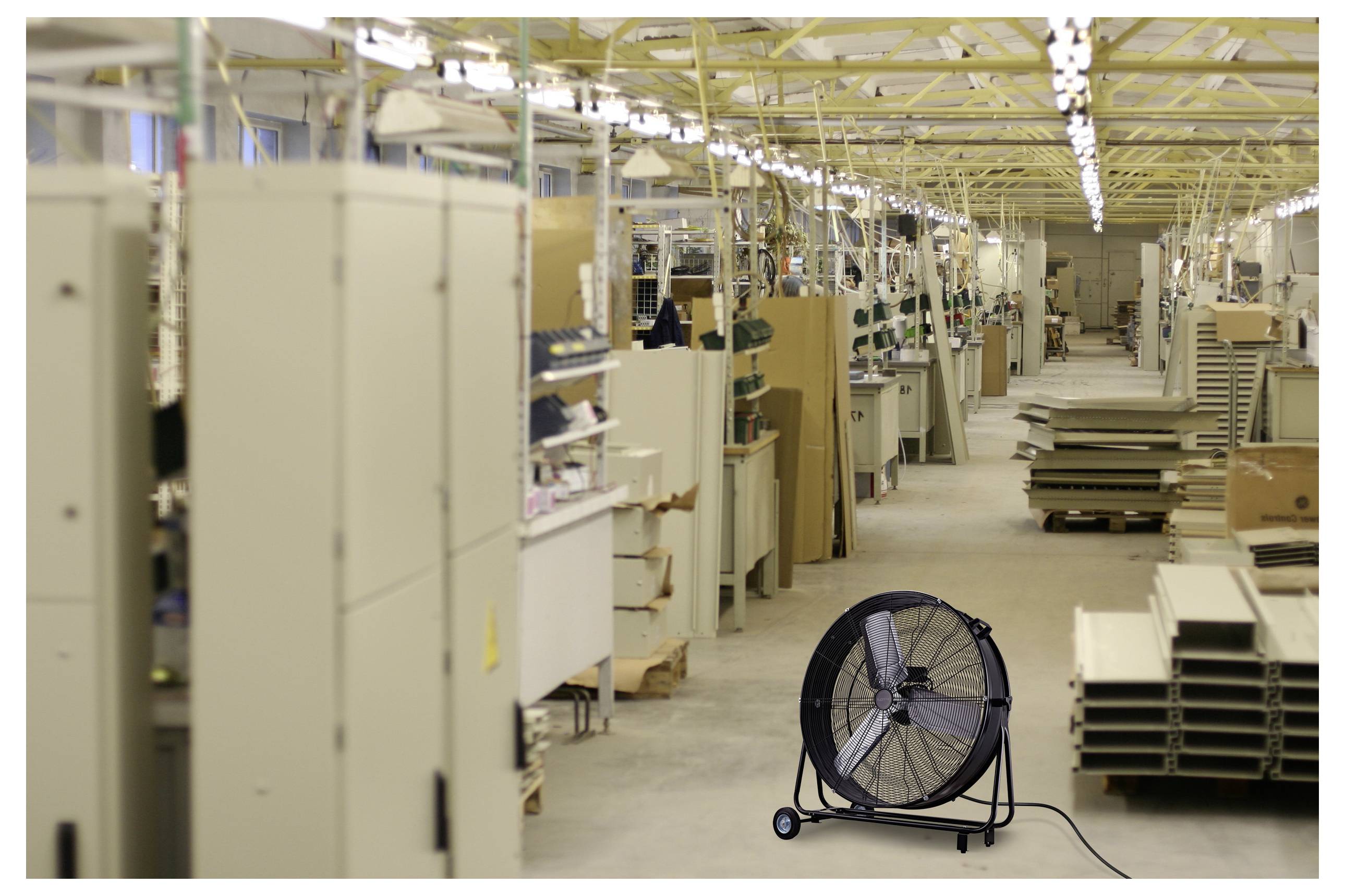 A large industrial fan is positioned on the floor of a spacious factory, surrounded by tall machinery and shelves filled with materials.
