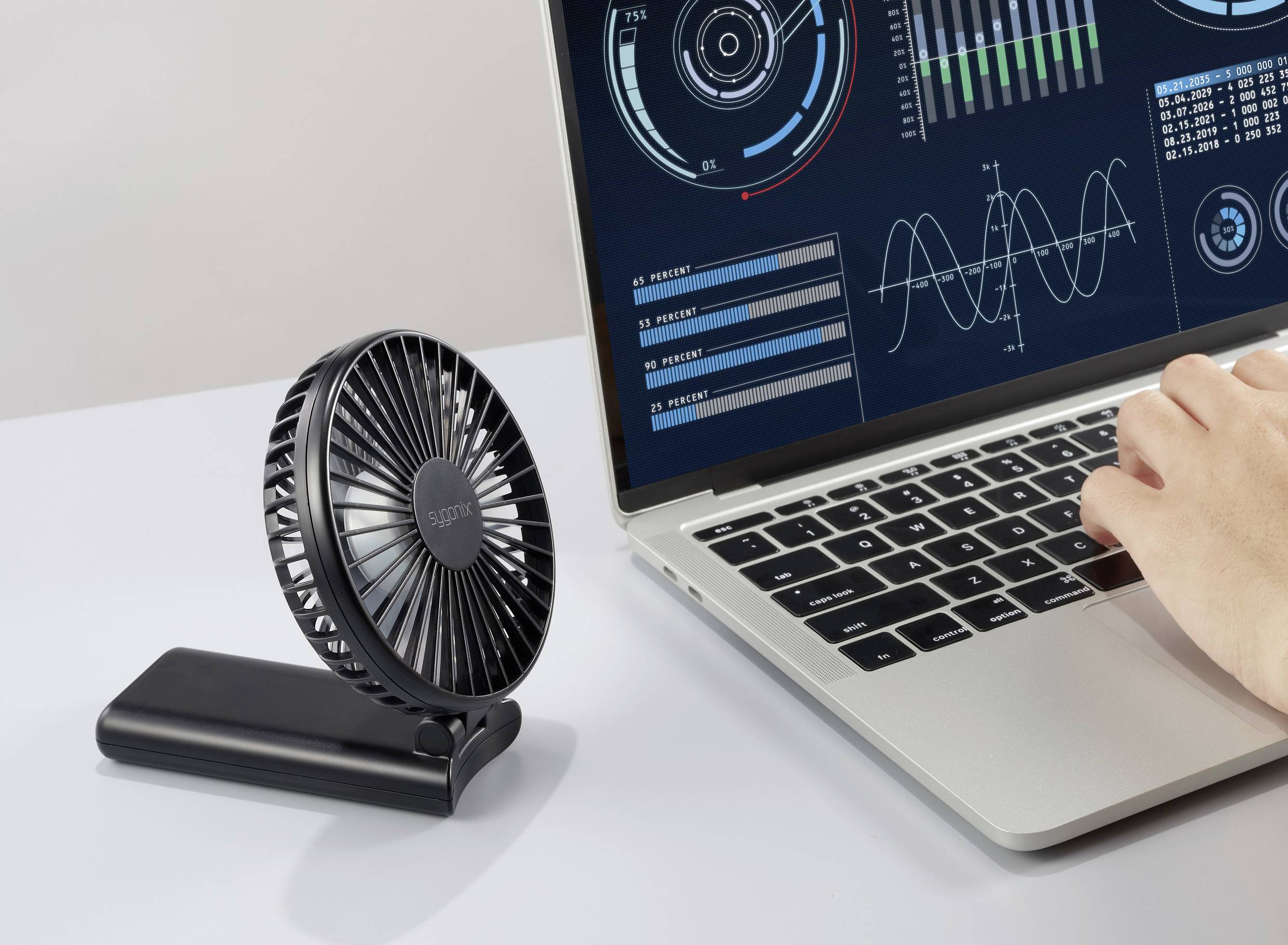 A portable fan positioned beside a laptop with analytical graphs on the screen; a hand typing on the laptop, suggesting a work setting.