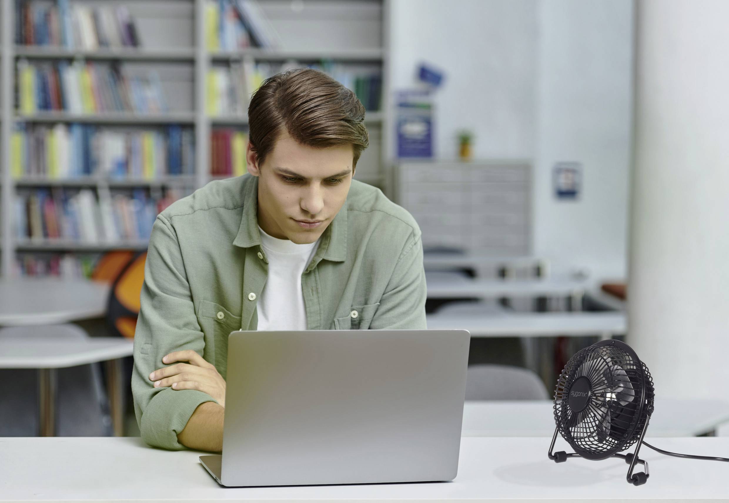A person is sitting at a table in a library, focused on a laptop. A small fan is on the table beside them, providing airflow.