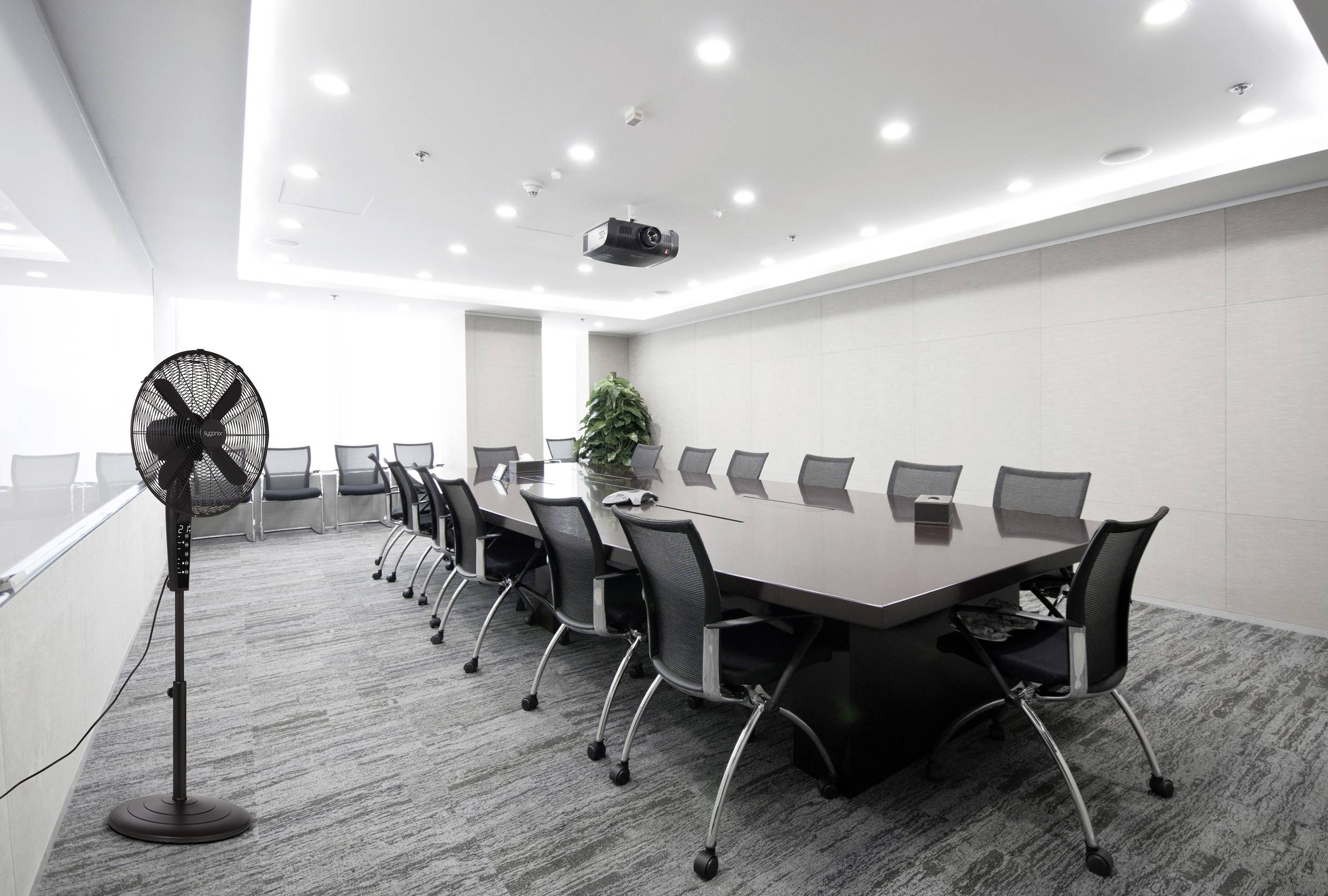 A modern conference room with a large rectangular table, multiple chairs, a projector on the ceiling, and a standing fan in the corner.