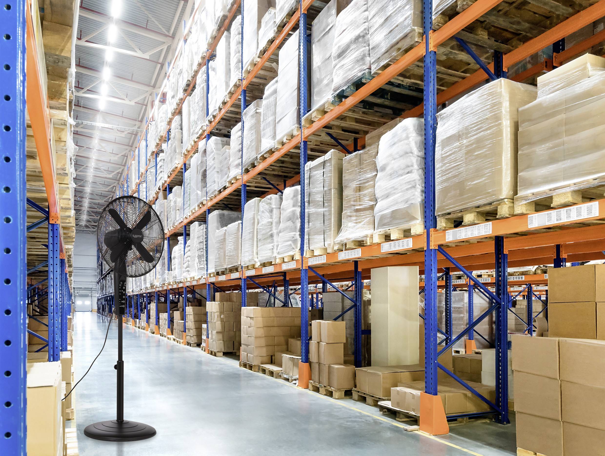 A large warehouse with tall, stocked shelves containing boxed goods. A standing fan is placed on the polished concrete floor near the foreground.