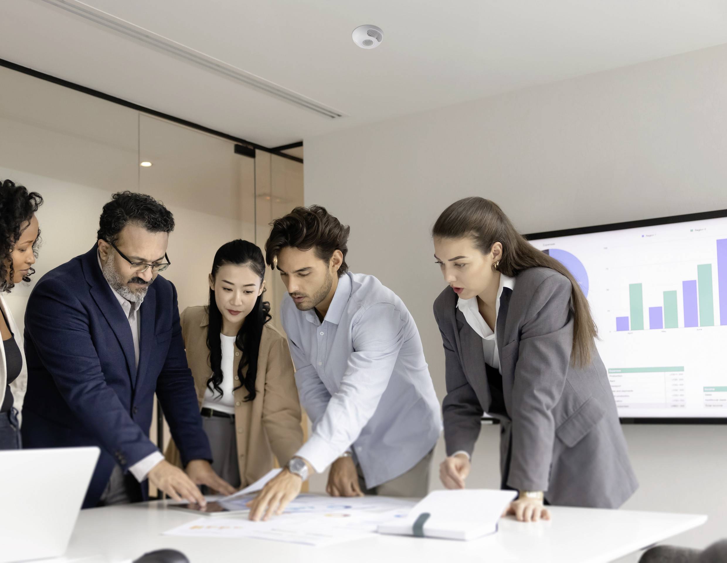 Colleagues in a meeting room review printed charts on a table. A screen displays a bar graph, suggesting data analysis or business discussion.