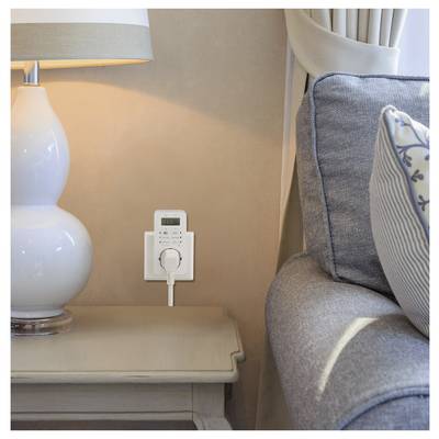 A digital plug timer is connected to a wall outlet beside a lamp and a sofa, indicating its use for efficient power management at home.