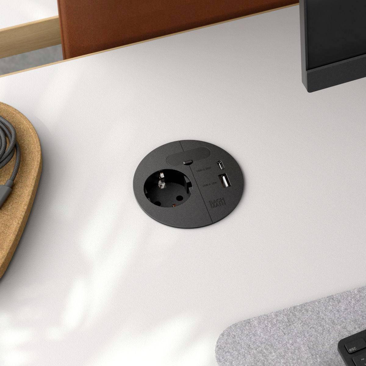 A power outlet and USB port embedded in a desk, with a monitor positioned next to it, suggesting a workspace setup.