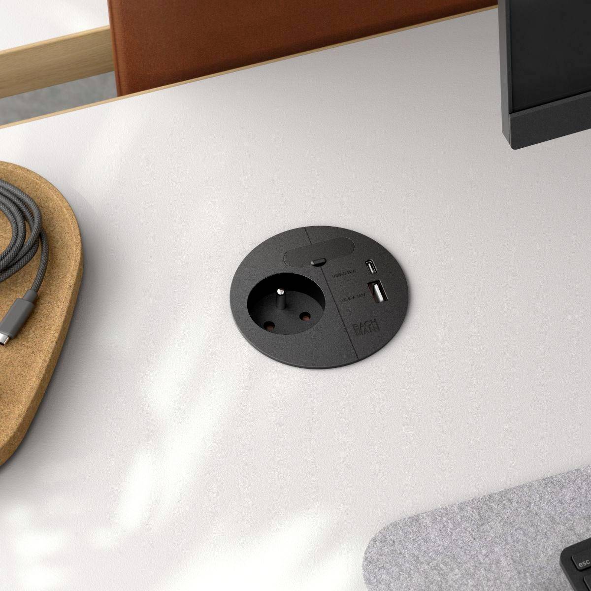 A round desk installed power hub with an electrical outlet and a USB port next to a leather chair and a gray felt mat, in an office setting.