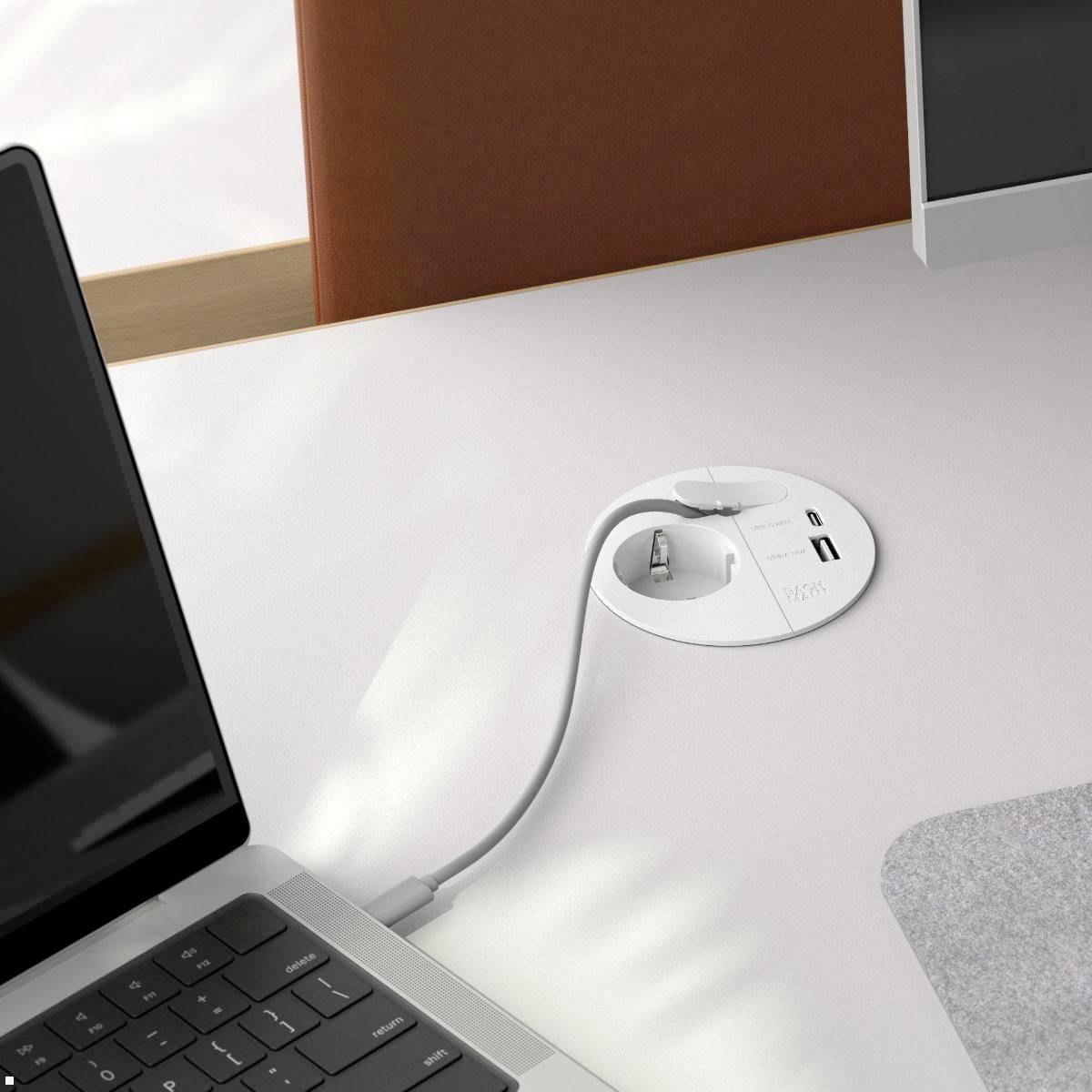 A desk features an integrated power outlet with USB and standard plug sockets. A charging cable is connected. A laptop is nearby.