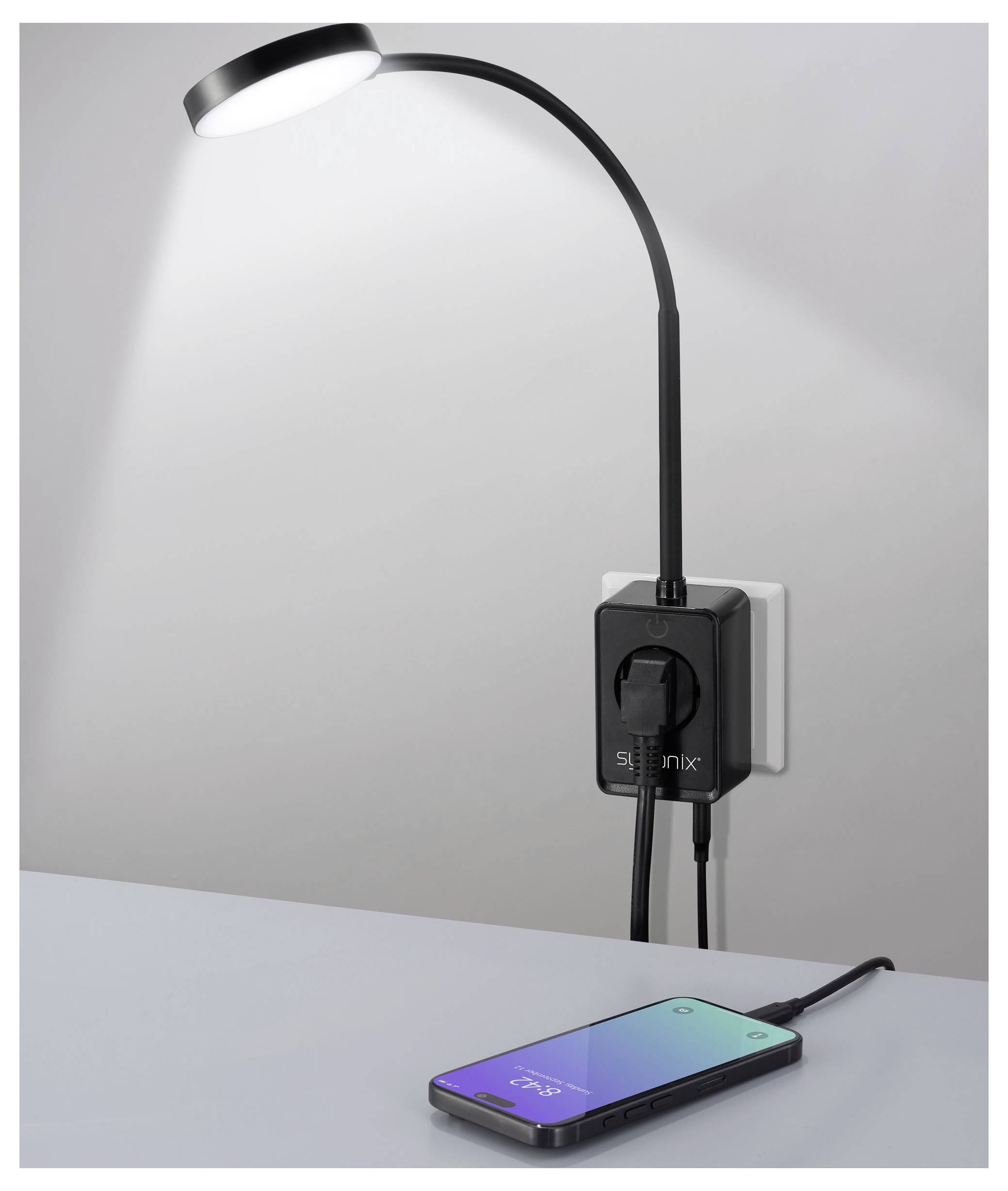 A flexible black lamp, plugged into a wall socket, illuminates a charging smartphone laying on a table with a colorful screen.