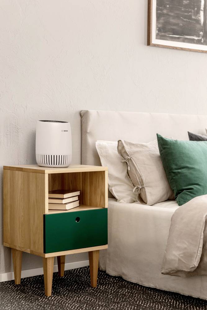 A neatly arranged bedroom with a wooden nightstand holding books, a small white air purifier, next to a bed with green and beige pillows.