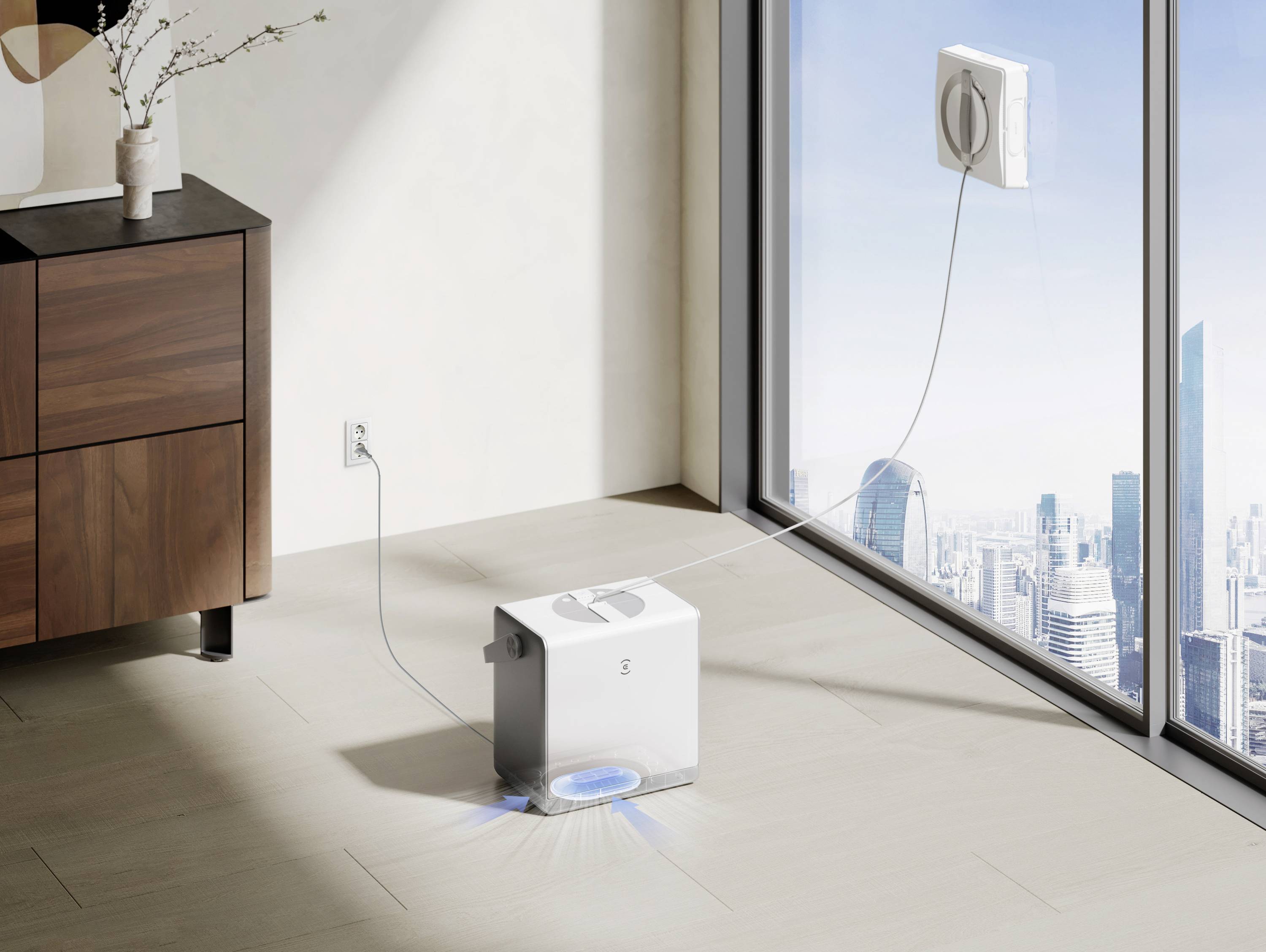A window-cleaning robot is attached and operating on a large glass window, powered by a cord connected to an indoor power outlet.