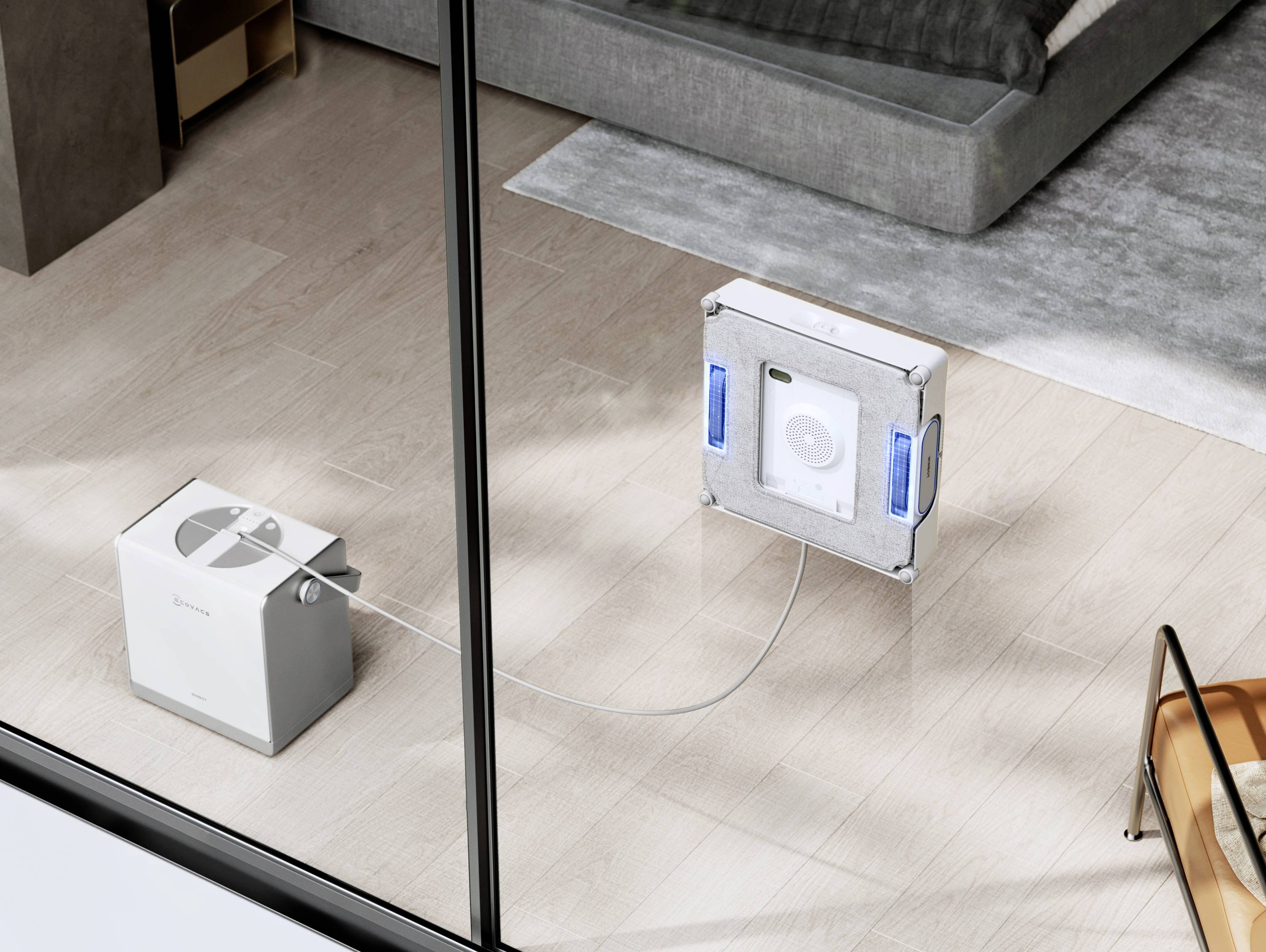 A robotic window cleaner is attached to a large glass window. It is connected by a cord to a base unit placed on the floor nearby.