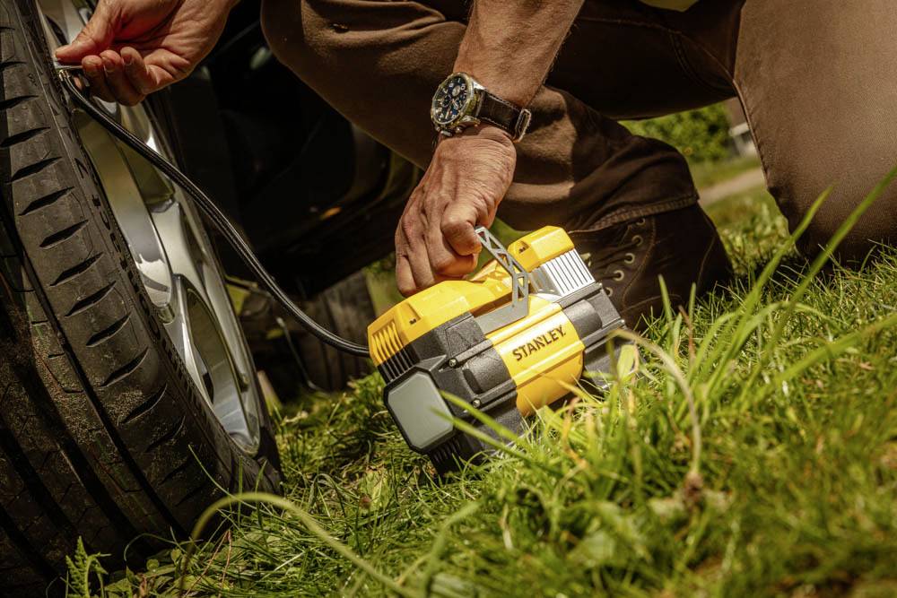 A person uses a portable air compressor to inflate a car tire, kneeling on grass, with the device labeled 'Stanley'.
