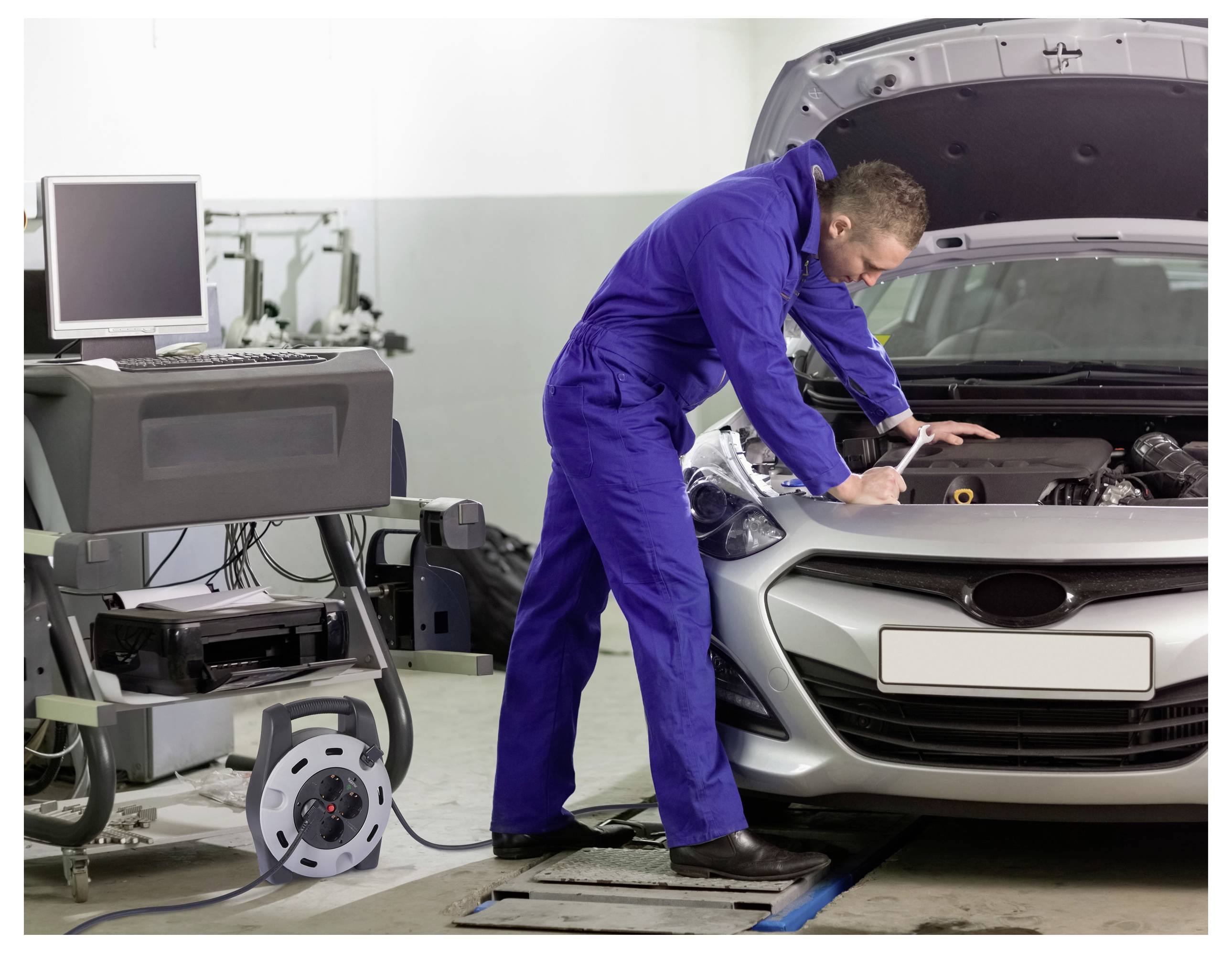 A mechanic in a blue jumpsuit checks a car engine in a garage, using diagnostic equipment connected to a nearby computer.