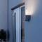 A modern outdoor wall light fixtures a sleek design, illuminating a gray building exterior next to a glass door, adding elegance and safety.