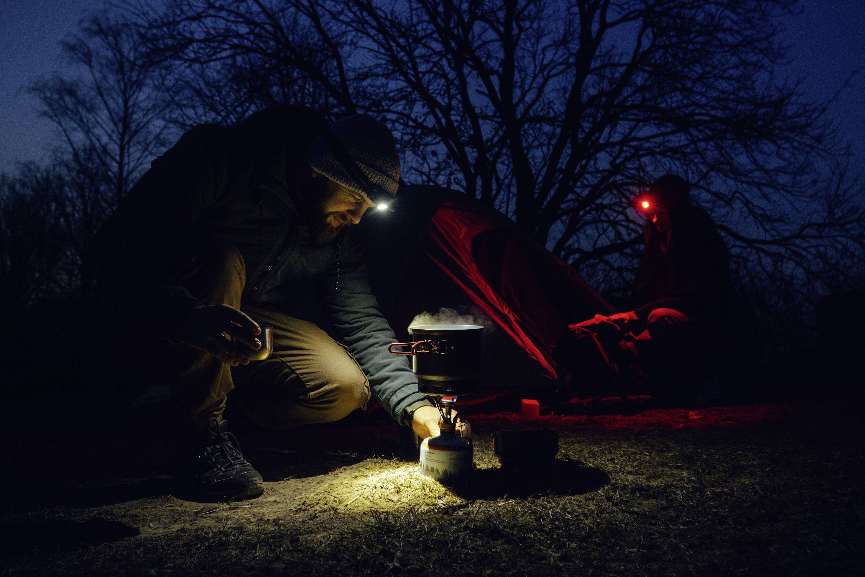 A person cooks on a portable stove beside a tent at night, wearing a headlamp; another person is seated nearby under a red light.