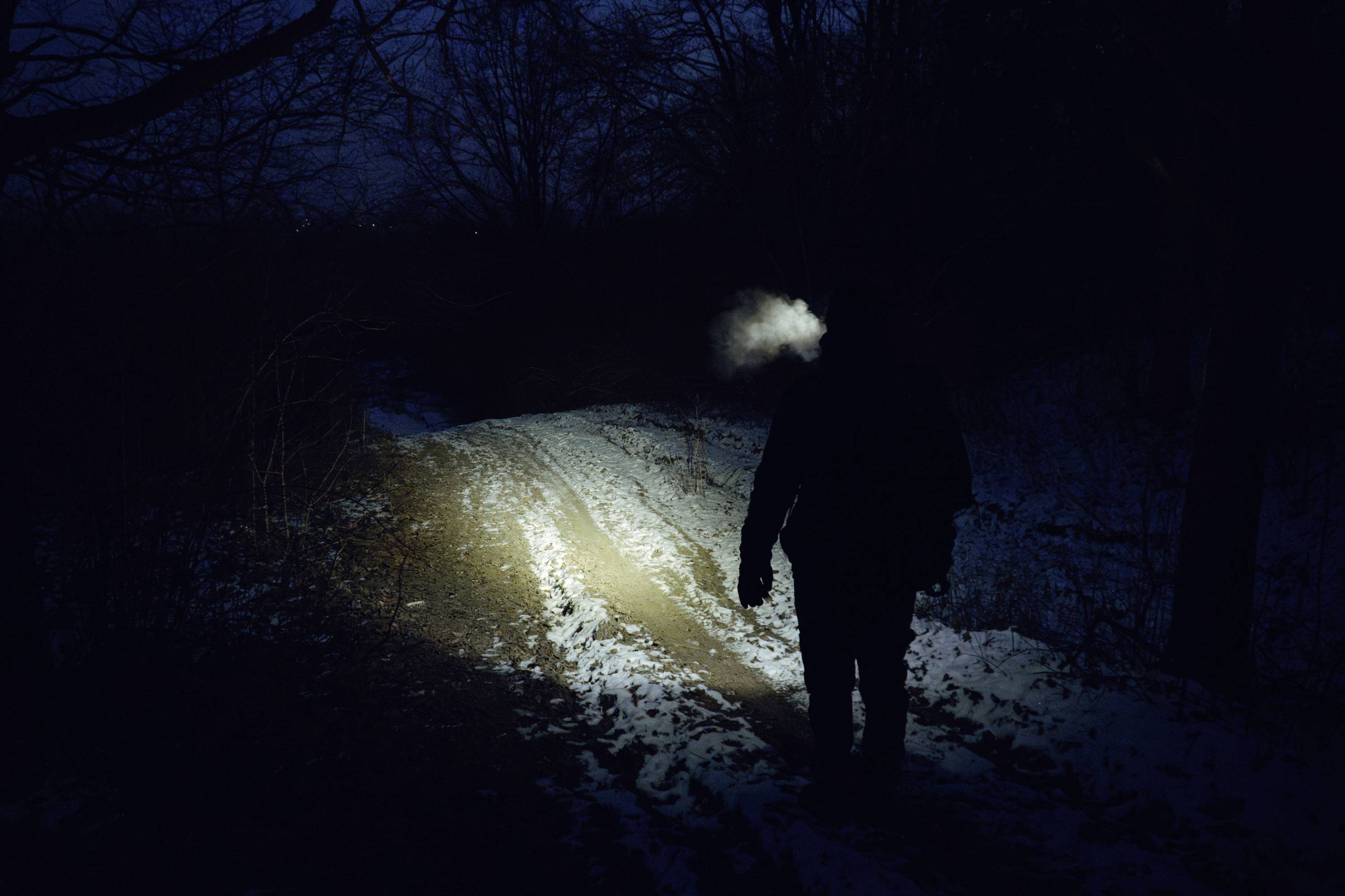 A person walks along a dimly lit, snowy path in a forest at night. Their breath is visible in the cold air, creating a mist.