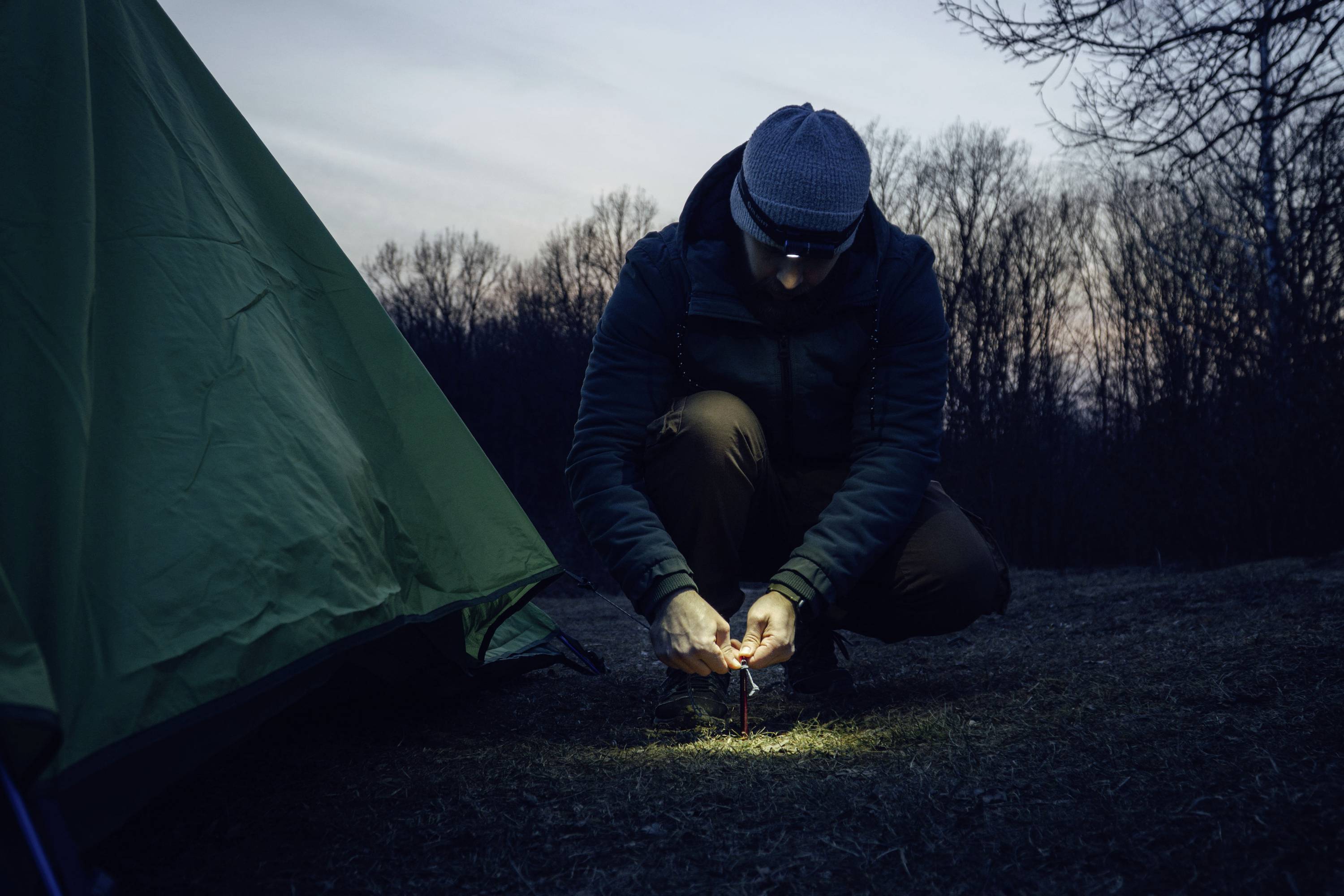 A person squats next to a green tent in a wooded area at dusk, concentrating on securing a tent stake into the ground.
