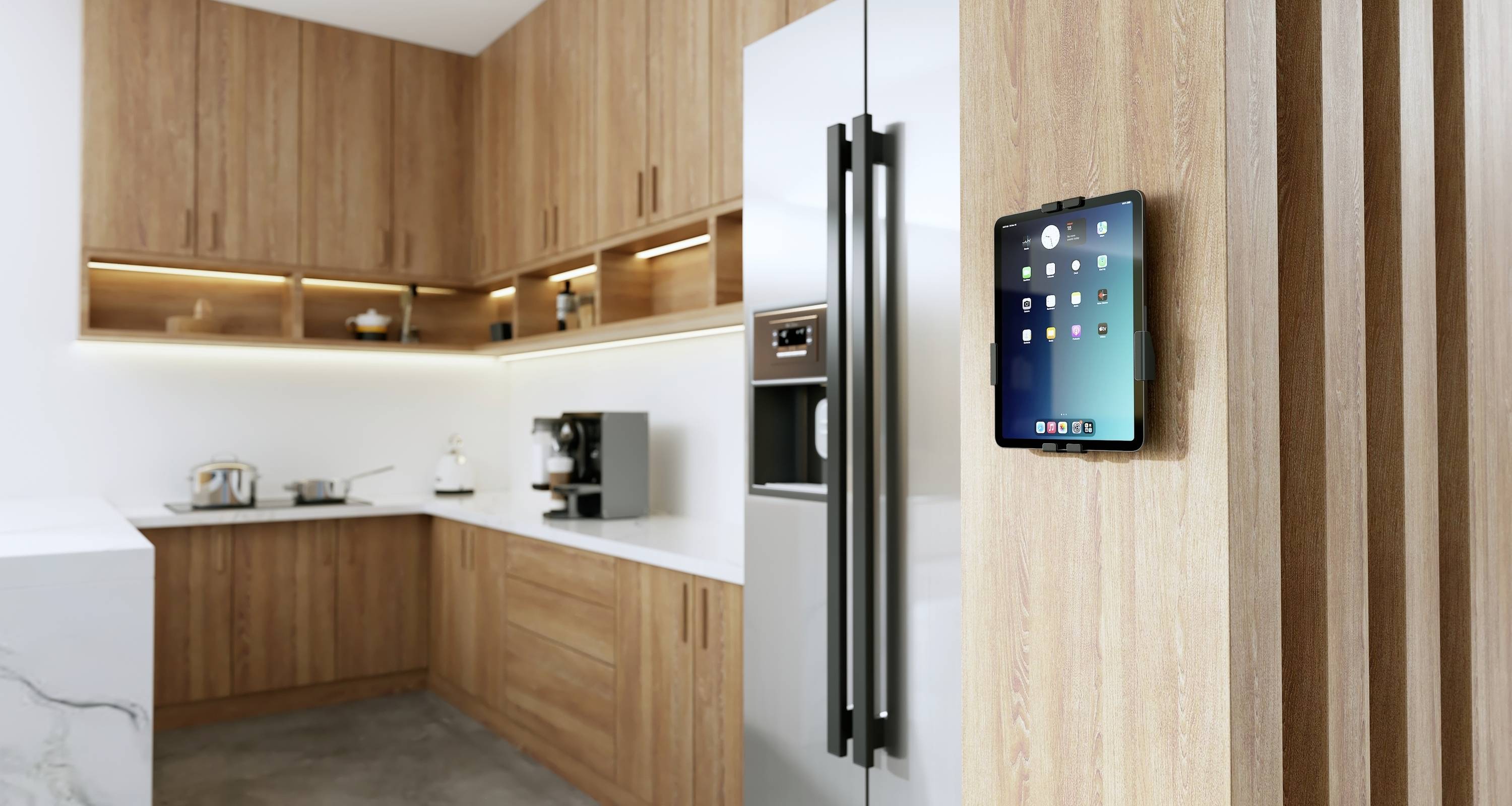 A modern kitchen with wooden cabinets features a tablet mounted on the wall, displaying a home automation interface.