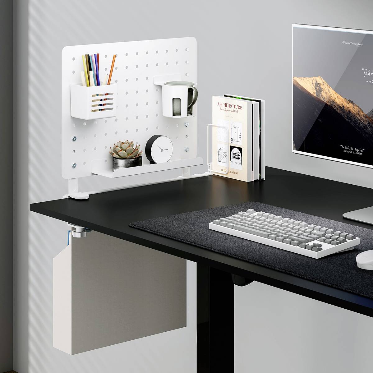 A modern workspace with a white pegboard holding stationery, a small succulent plant, clock, a tablet displaying mountain scenery, and a keyboard on a black desk.