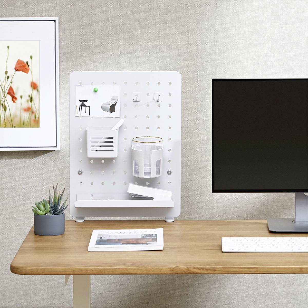 Modern desk setup with a beige corkboard, small potted plants, a computer with keyboard and mouse, and a framed floral artwork on the wall.