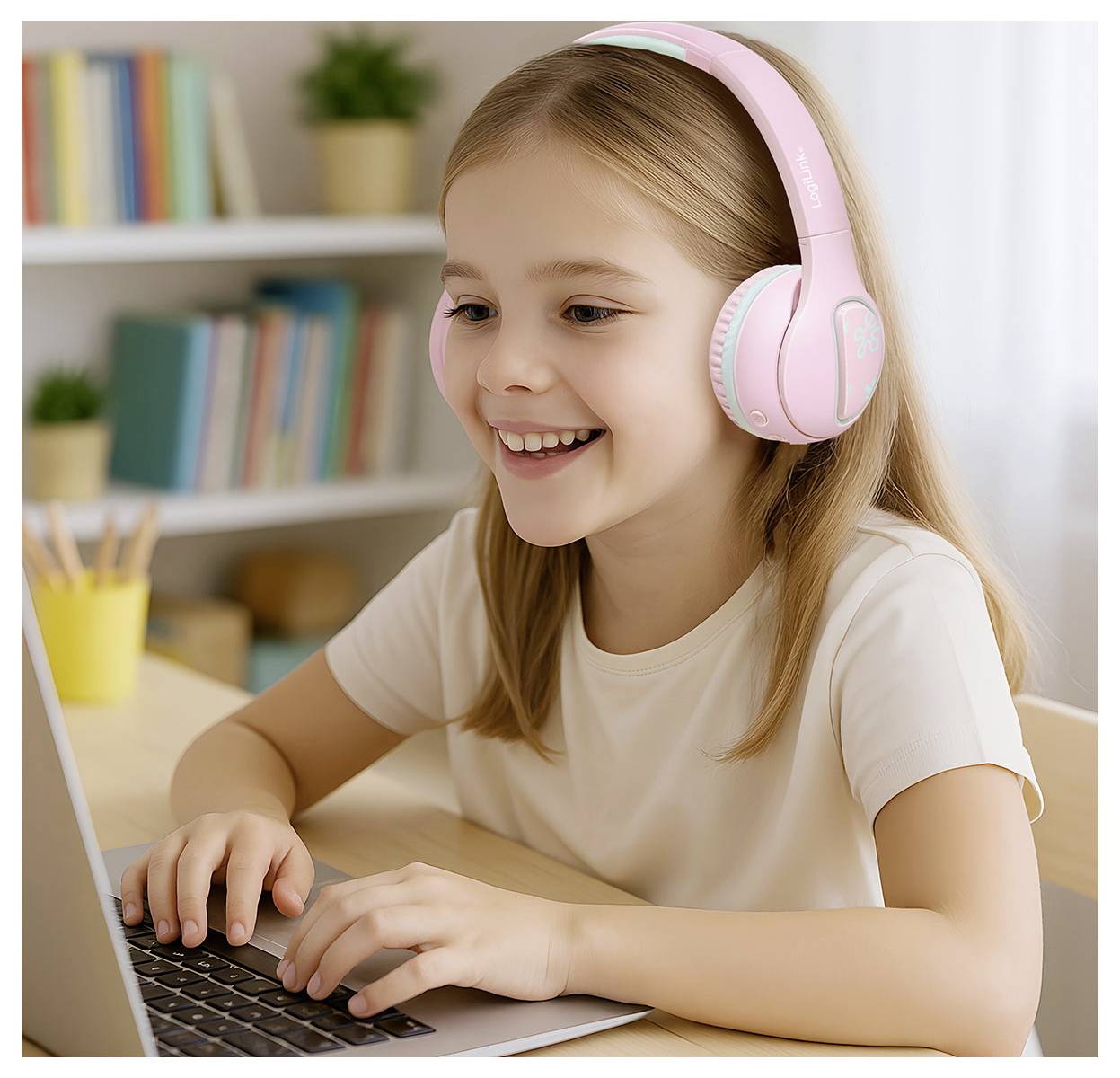 A smiling young girl with headphones uses a laptop at a desk in a room with bookshelves, indicating online learning or entertainment.