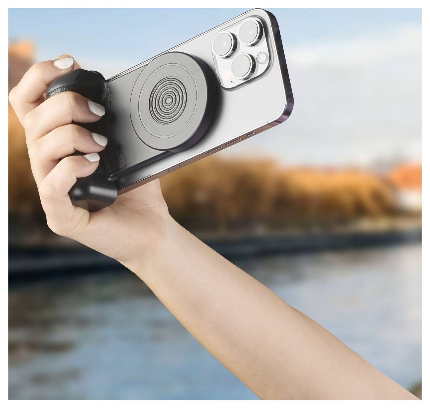 A hand holding a smartphone with a circular grip attachment on the back, set against a blurred outdoor backdrop near water.