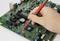 A hand uses a red probe on a computer motherboard, testing electronic components.