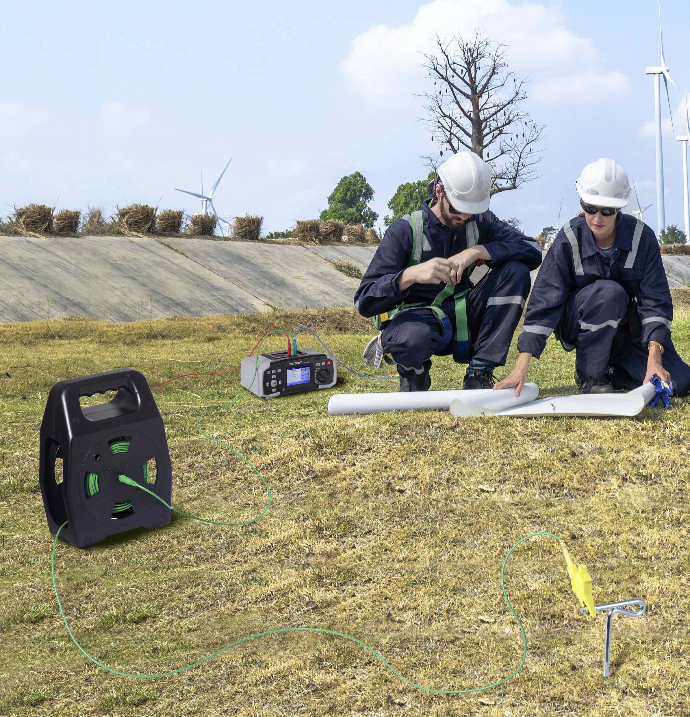 Two workers in helmets and safety gear discuss plans on a grassy field with wind turbines. Equipment lies nearby, including a generator.