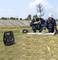 Two workers in helmets and safety gear discuss plans on a grassy field with wind turbines. Equipment lies nearby, including a generator.